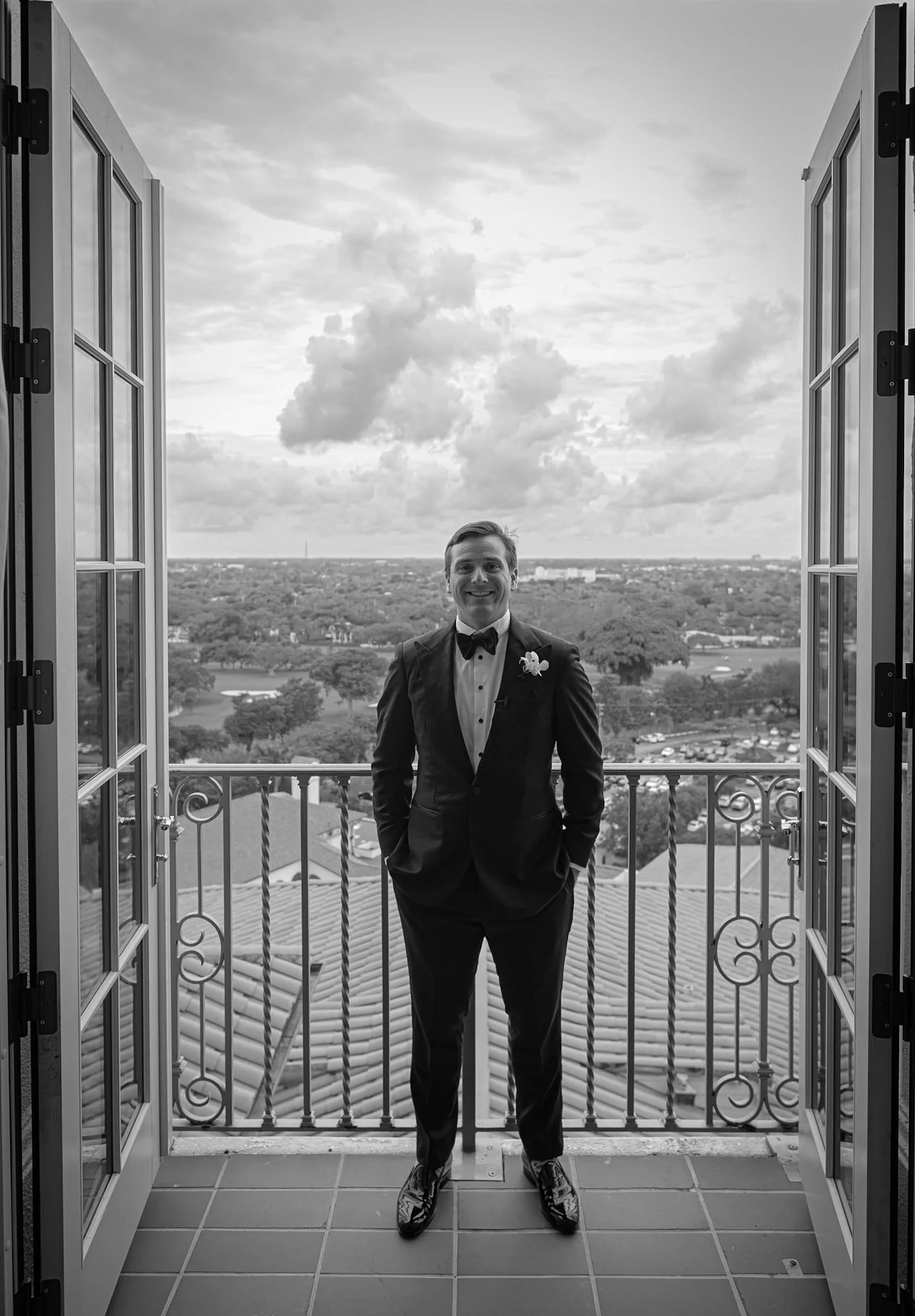 A man in a tuxedo standing on a balcony with an open window behind him, overlooking a cityscape and cloudy sky.