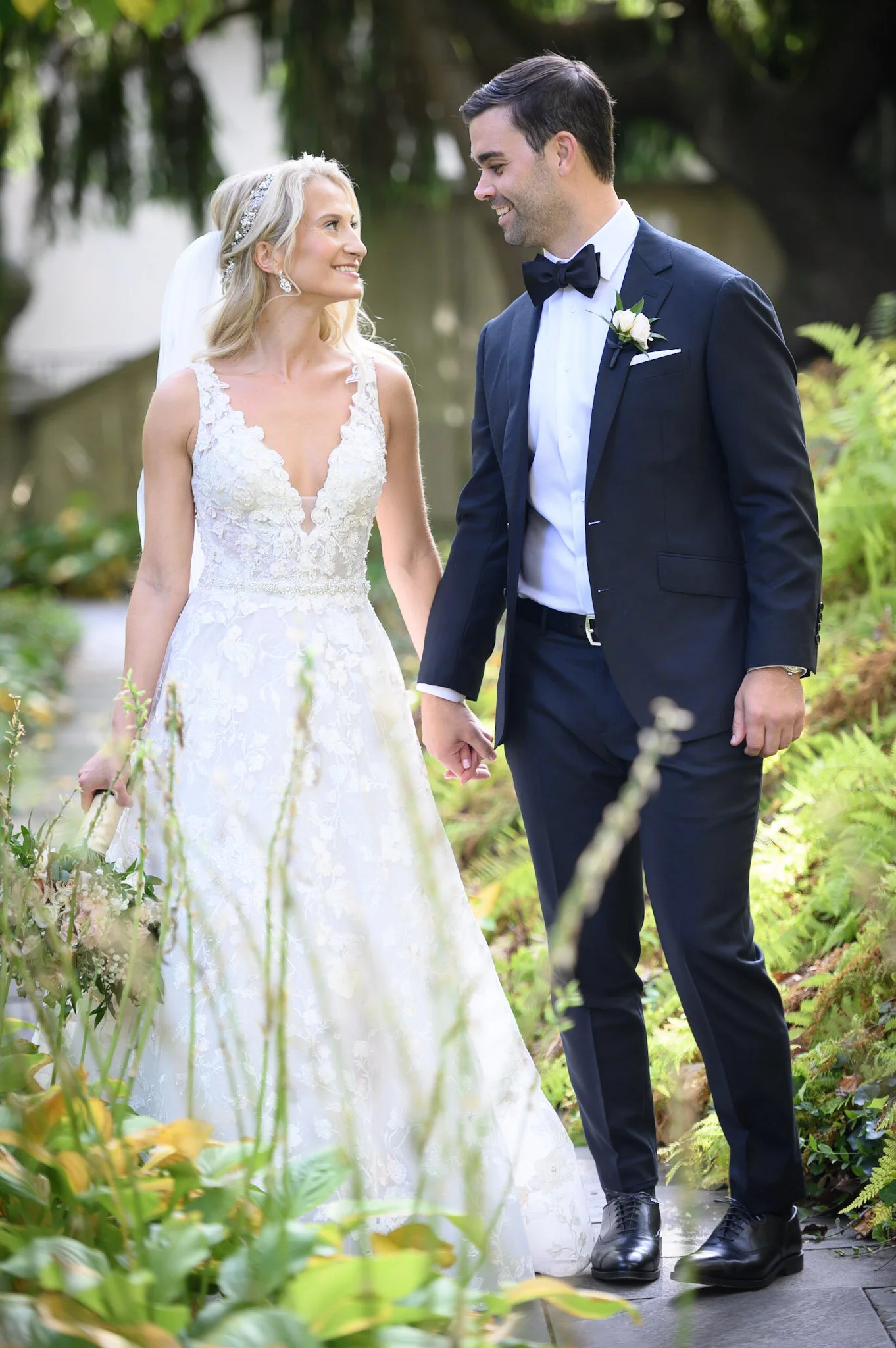 A bride and groom holding hands, standing outdoors on a garden pathway, smiling and looking at each other.