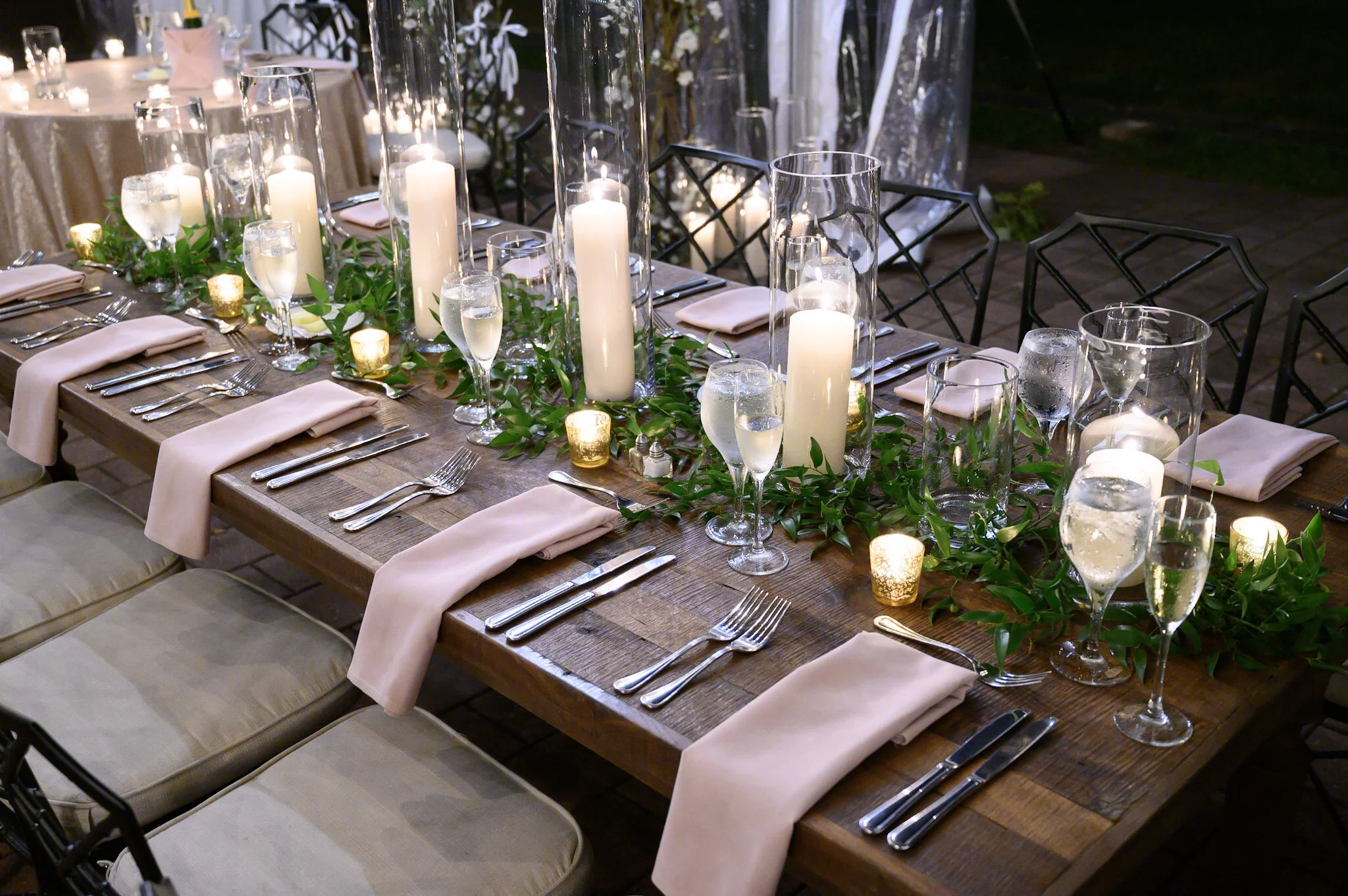 A long wooden dining table decorated with candles in glass and metal holders, greenery, napkins, and silverware, set up for an outdoor evening event.