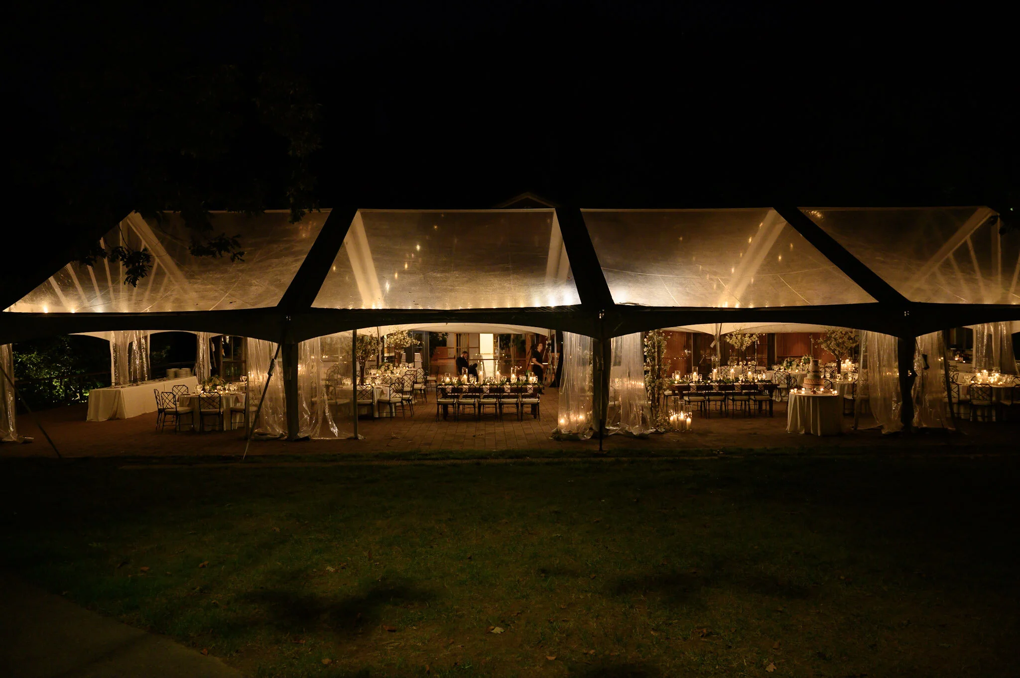 Night view of an outdoor wedding or event reception under a transparent tent with tables, chairs, and candlelit decorations inside, surrounded by dark trees and grass.