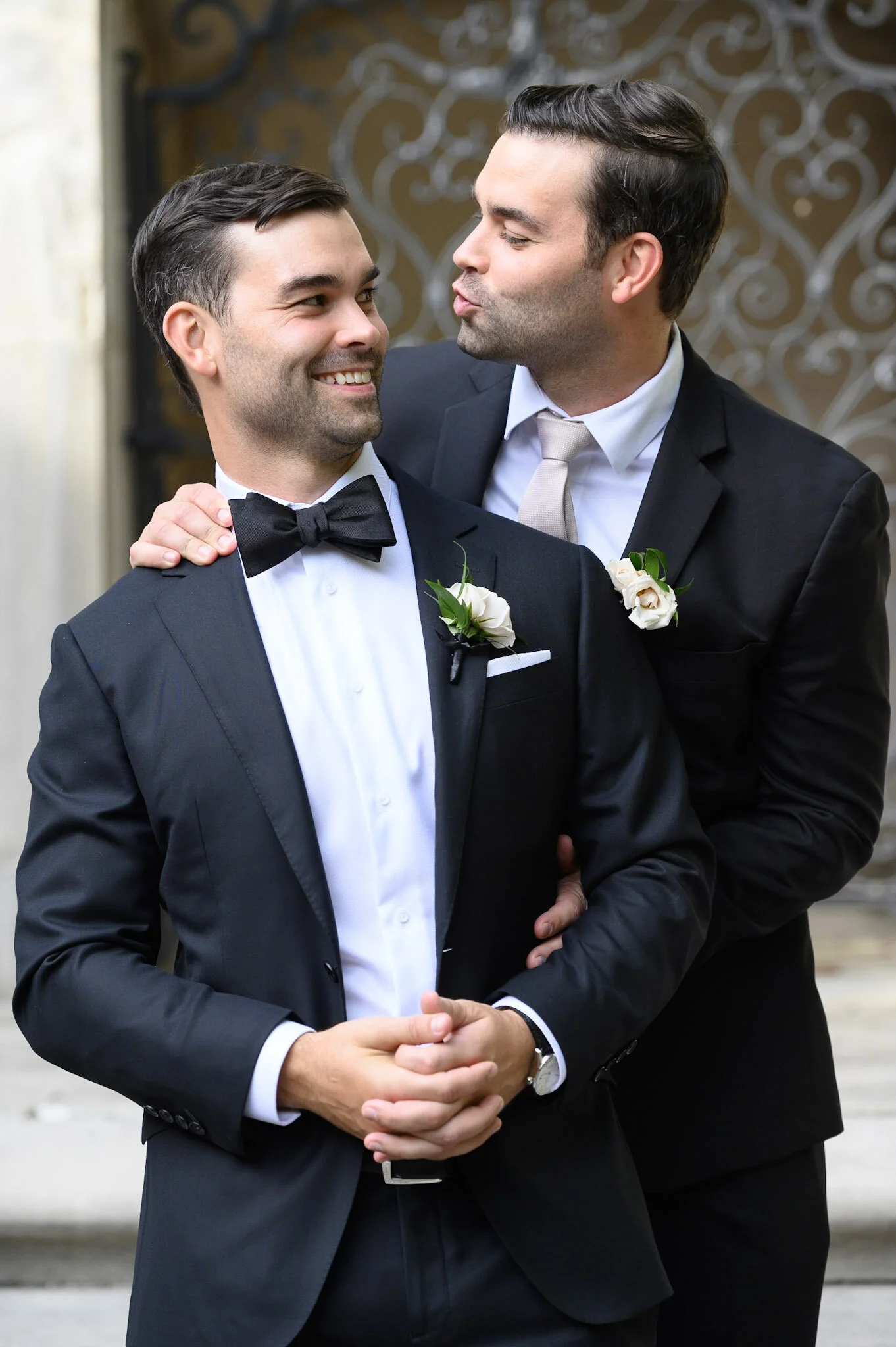 Two men in tuxedos greet each other with a hug, smiling warmly. The man on the left is wearing a black tuxedo with a black bow tie, holding his hands clasped. The man on the right is wearing a black suit with a white shirt and a light-colored tie, wi