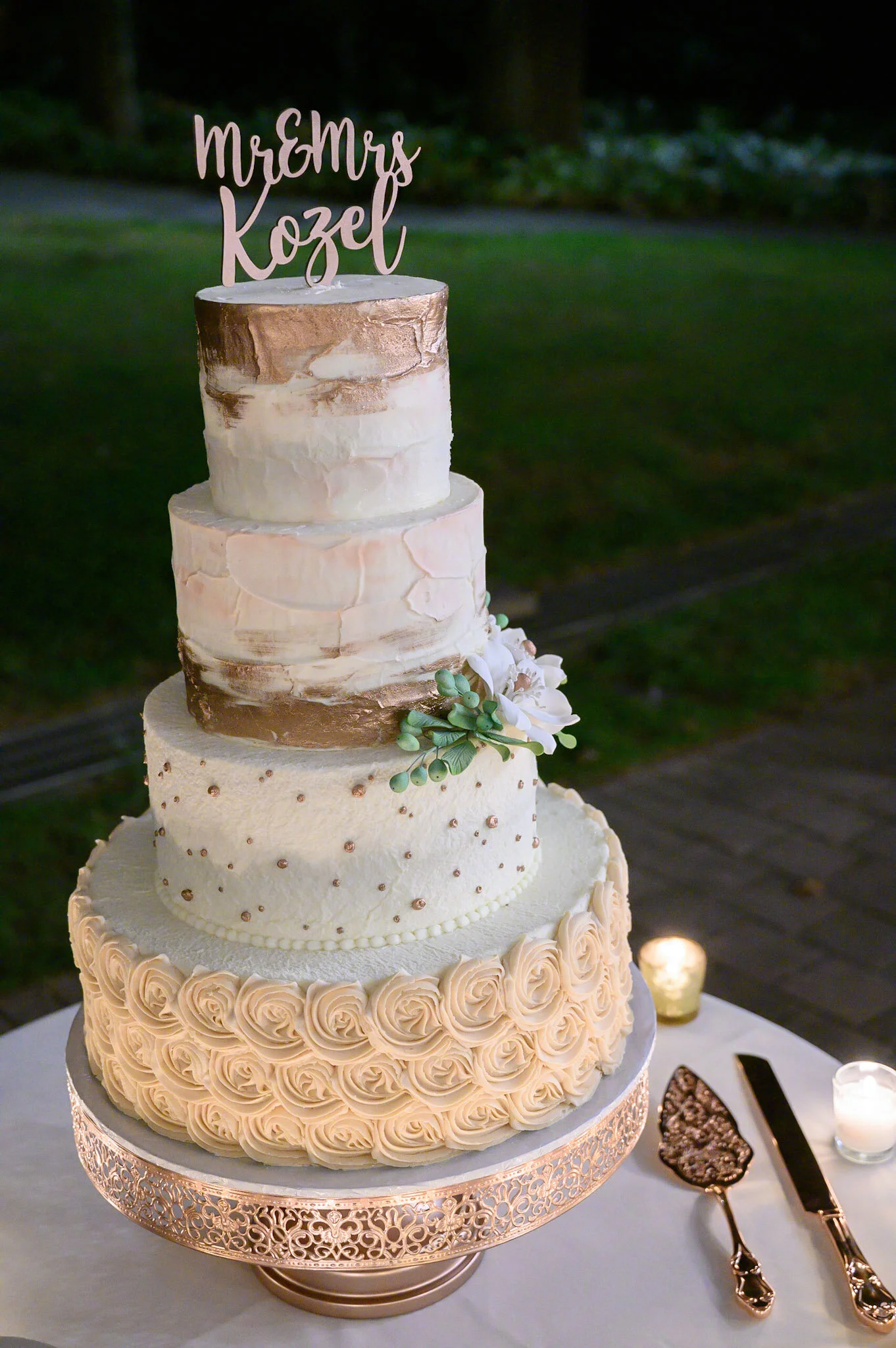A four-tier wedding cake decorated with roses, rosette piping, and gold accents, topped with a cake topper that reads 'Mr & Mrs Kozel' in pink script. The cake sits on a gold ornate cake stand, with a cake knife and server on the table alongside cand