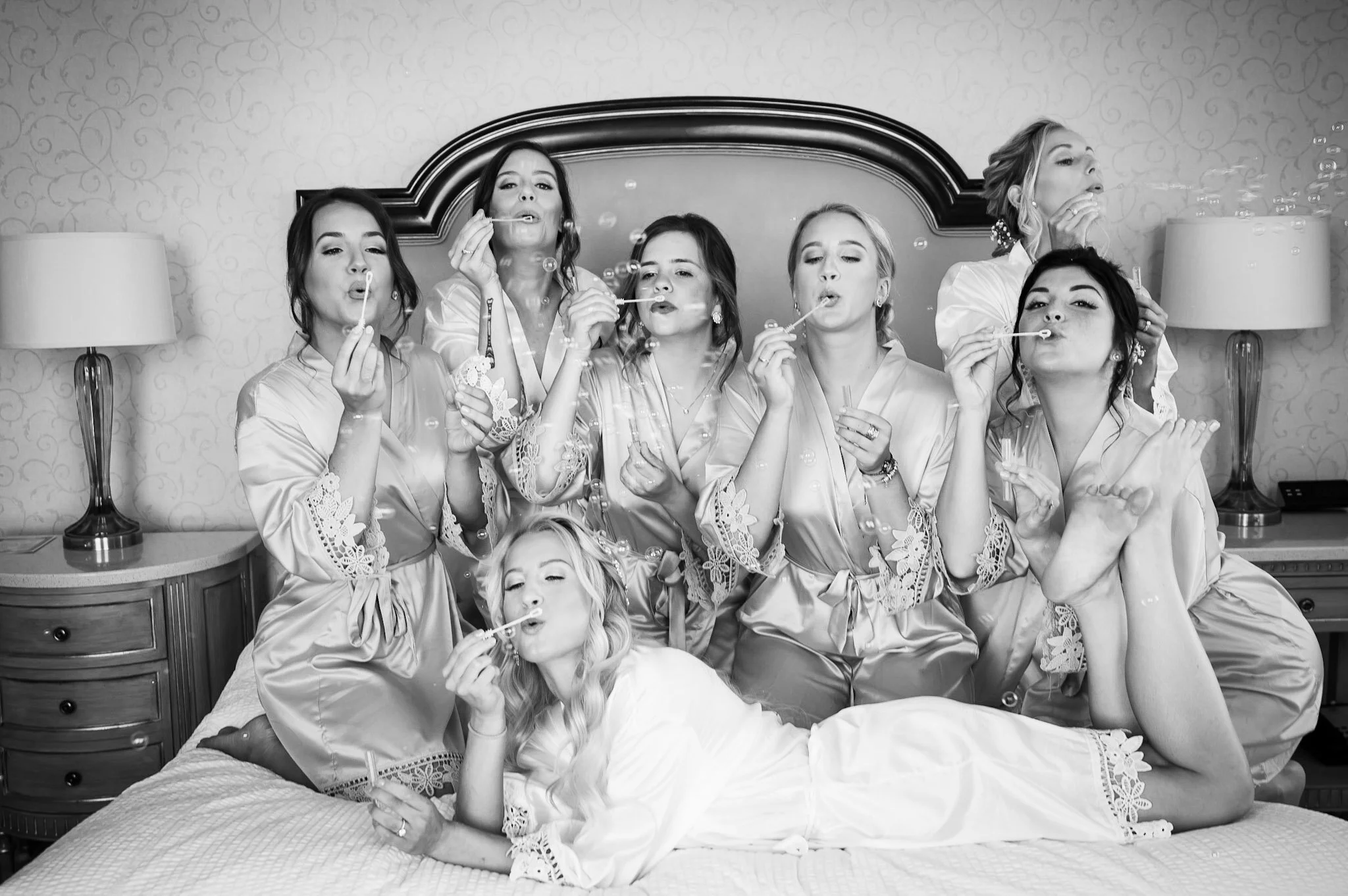 A group of women in satin robes sitting and lying on a bed in a hotel room, blowing bubbles with bubble wands, celebrating an occasion.