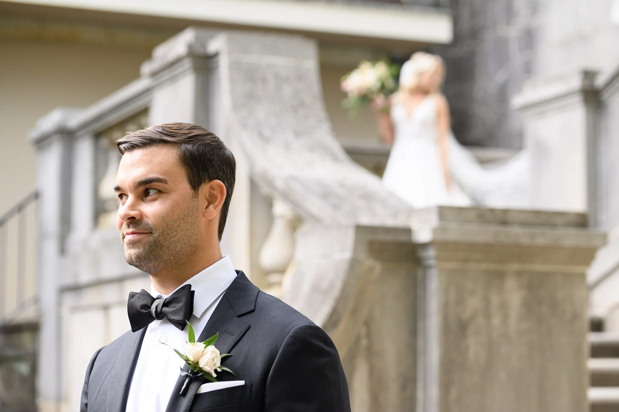 A groom with dark hair and a beard, dressed in a black tuxedo with a white shirt and black bow tie, stands in front of a stone staircase with a blurred bride in the background holding a bouquet of pink and white flowers.