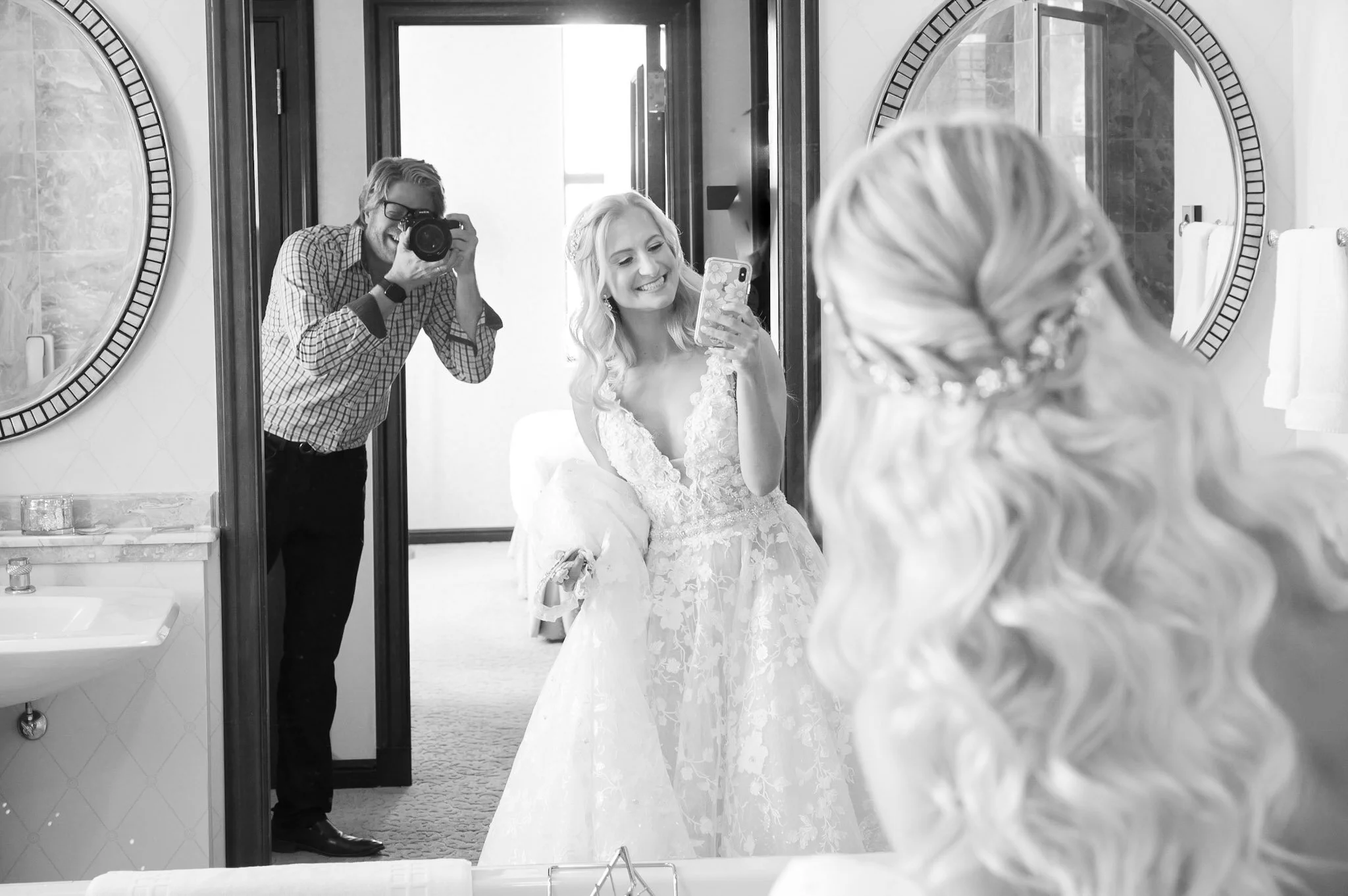 A bride taking a selfie in a mirror with a bridesmaid in front of her, while a man with glasses and a checkered shirt films them with a camera in a bright room.
