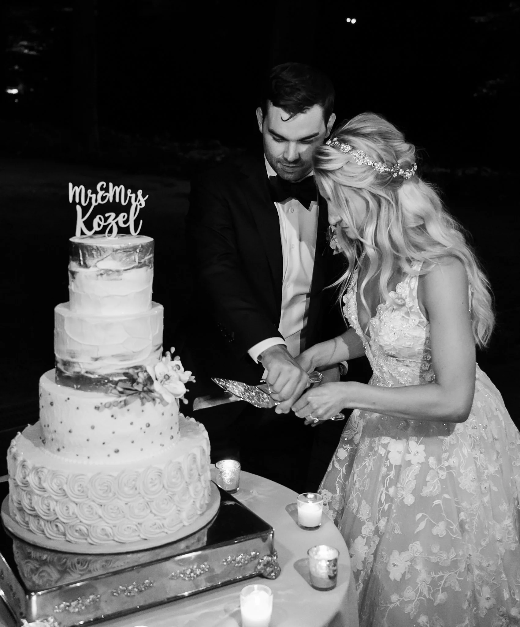 A bride and groom cutting a wedding cake together at their wedding reception. The cake is decorated with rose designs and has a topper that says 'Mr. & Mrs. Kozel'. There are candles on the table.