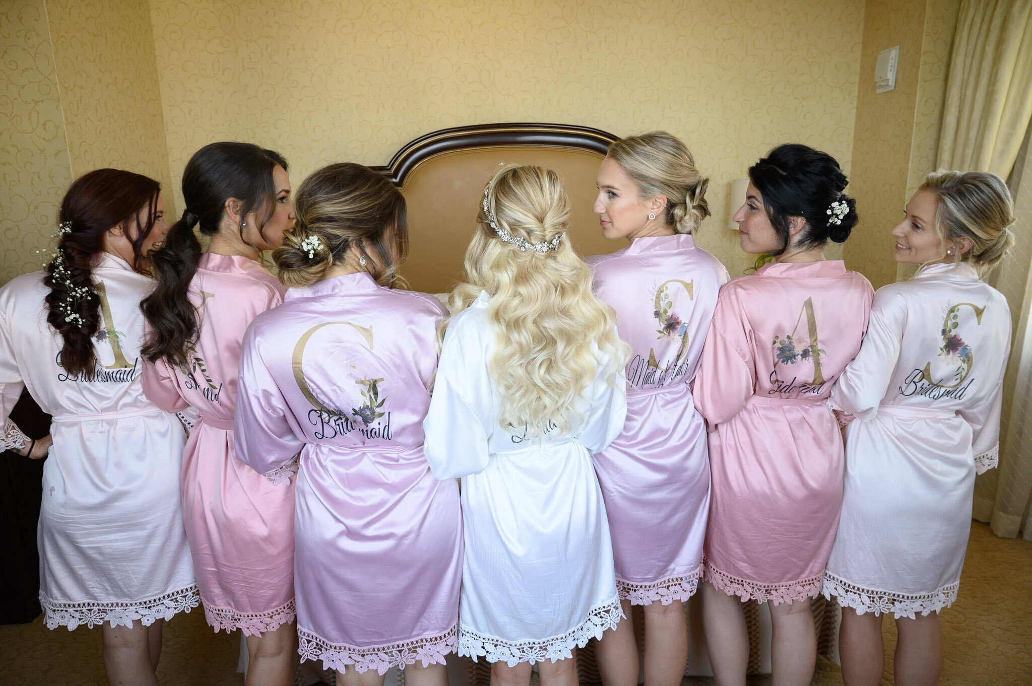 A bride and six bridesmaids wearing matching pink and white robes with floral embroidery, standing with their backs to the camera in a room with yellow wallpaper and a wooden headboard.