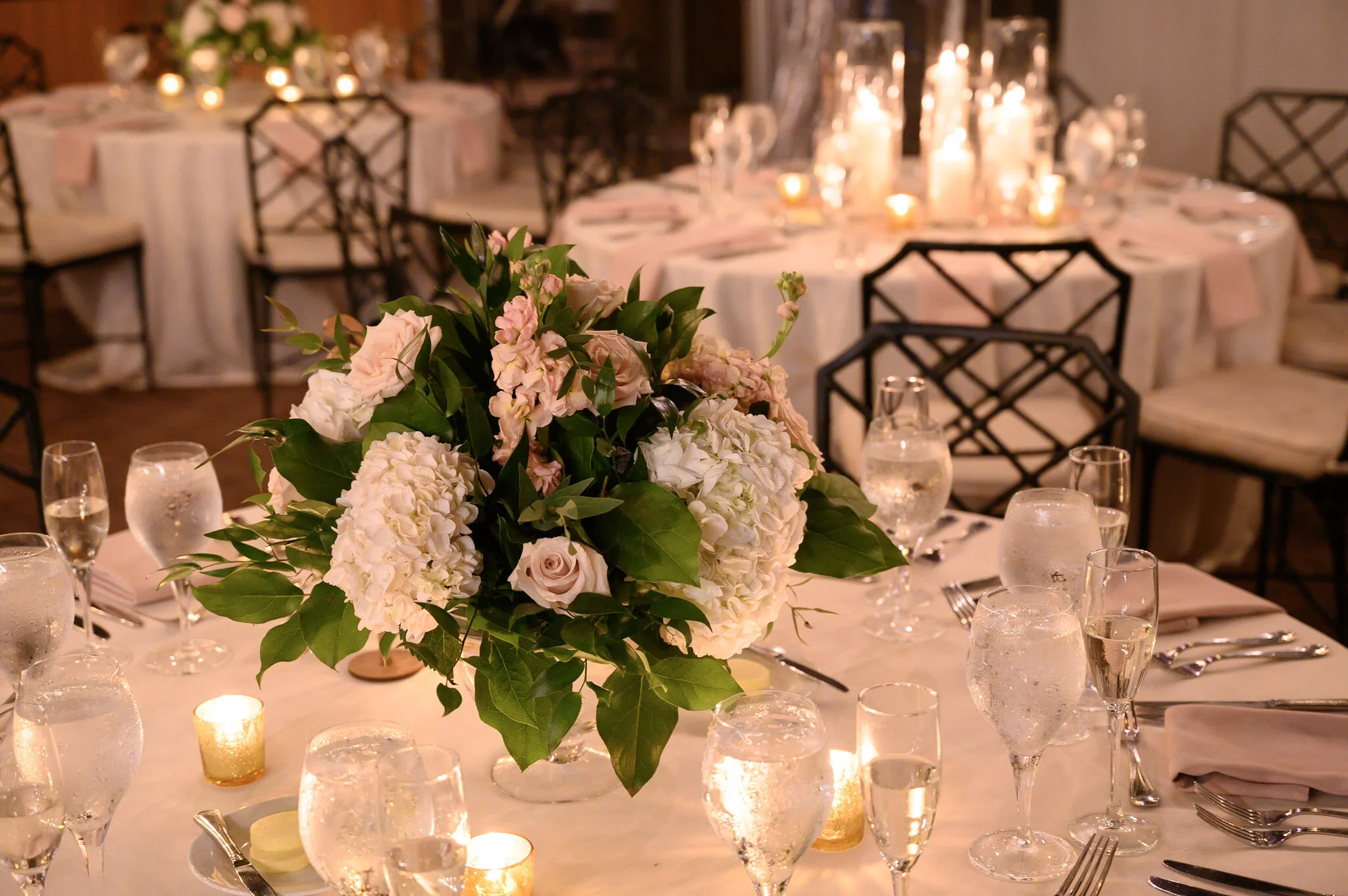 Elegant banquet table decorated with a large floral centerpiece of white and pink flowers with green foliage, surrounded by lit candles, glasses, and tableware in a warmly lit dining room.