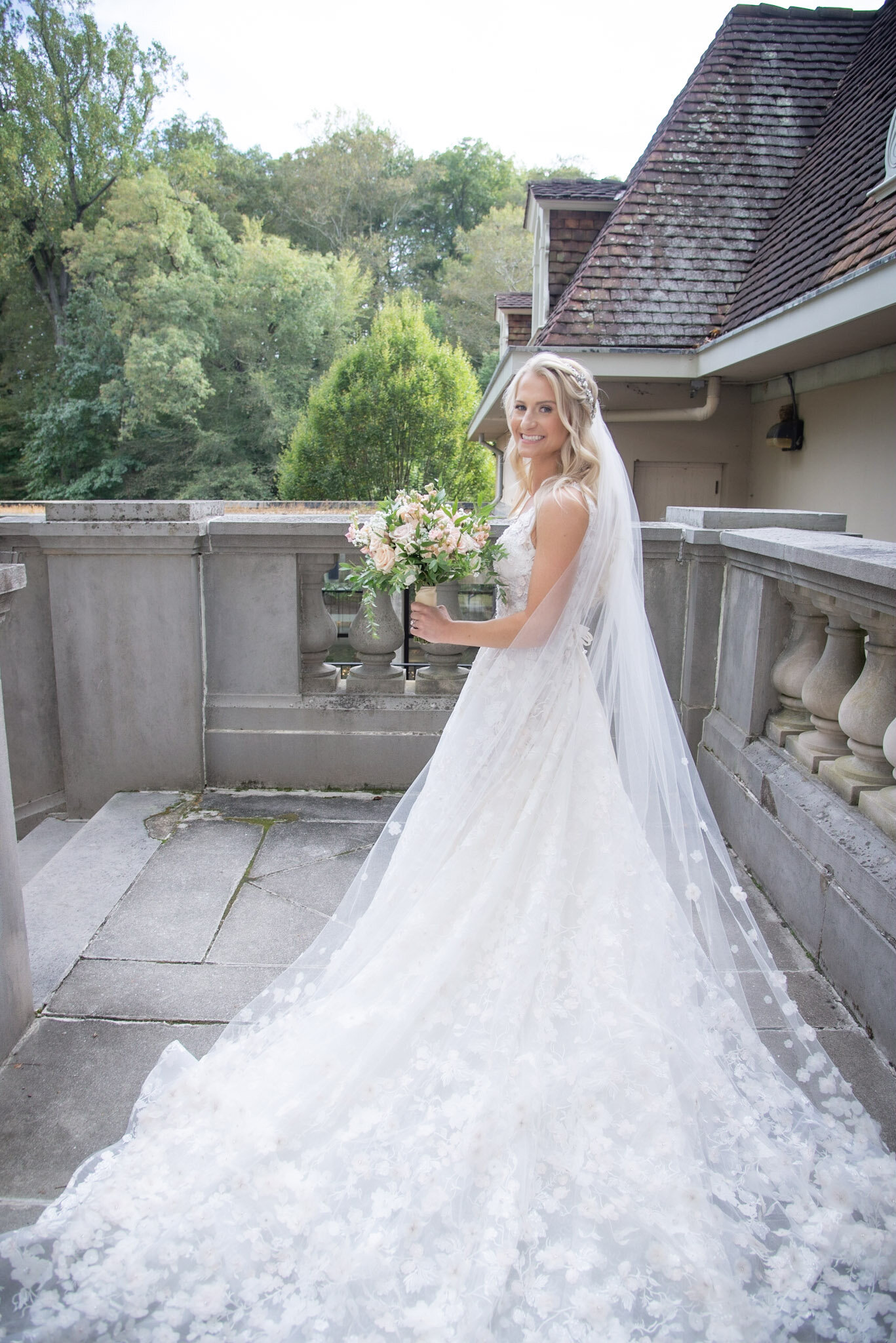 A bride standing on an outdoor stone terrace, holding a bouquet of pink and white flowers, wearing a white wedding gown and veil, with a background of green trees and a house with a tiled roof.