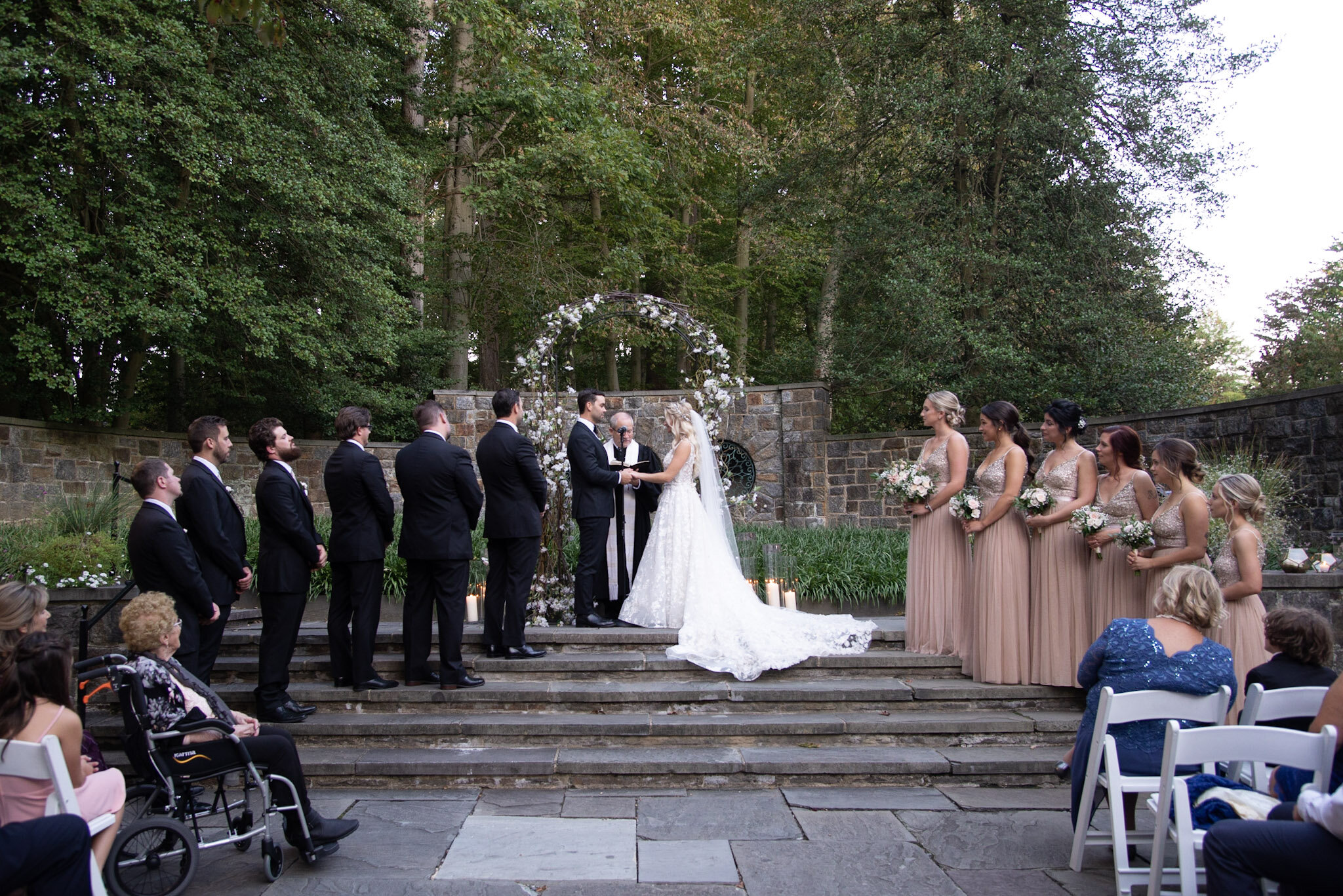 A wedding ceremony taking place outdoors on stone steps surrounded by trees, with a bride and groom exchanging vows, and bridesmaids and groomsmen standing on either side.
