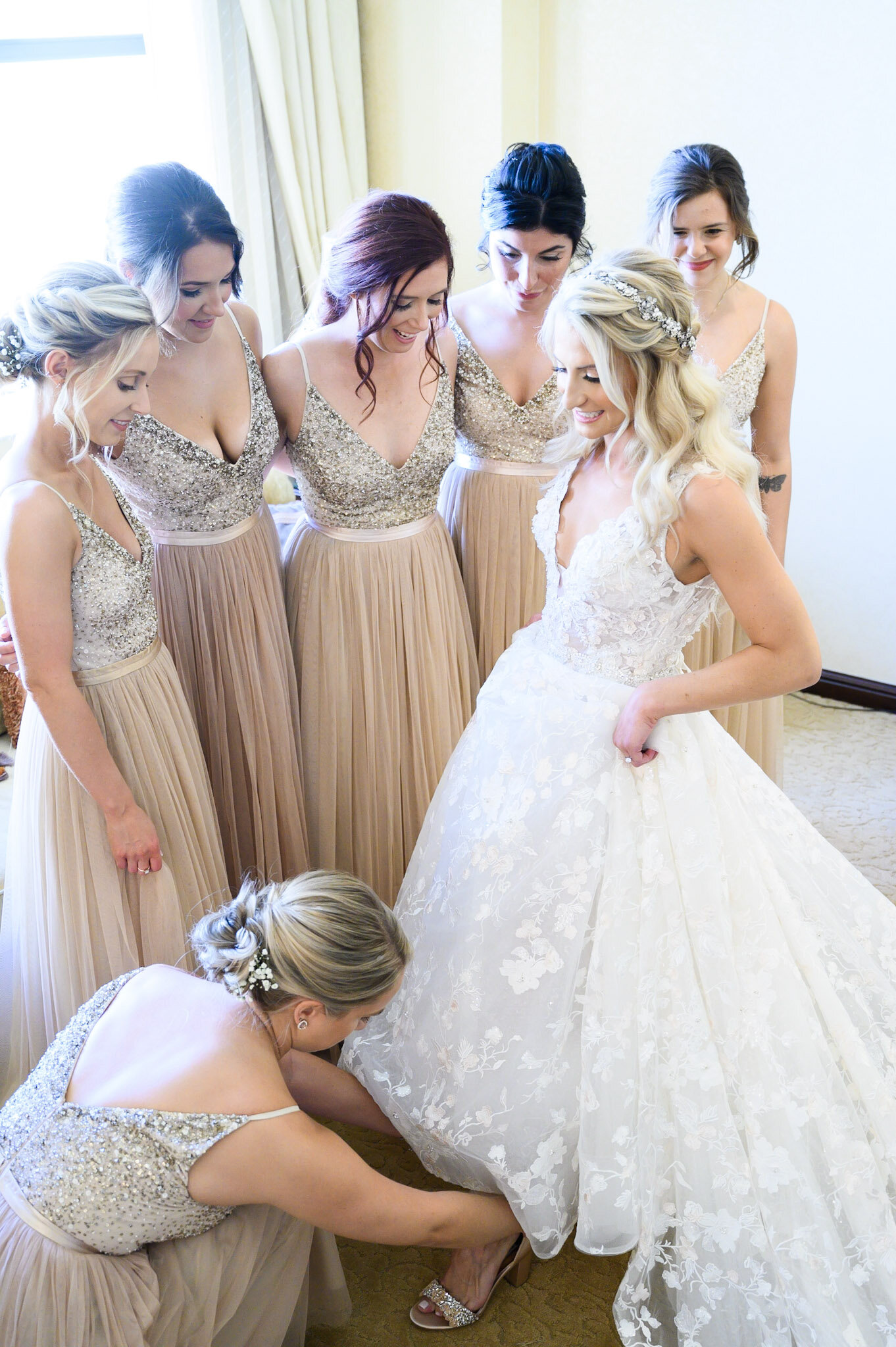 A group of women in matching beige and silver dresses gathered around a bride in a white wedding gown. One woman is adjusting the bride's dress while others look on happily.