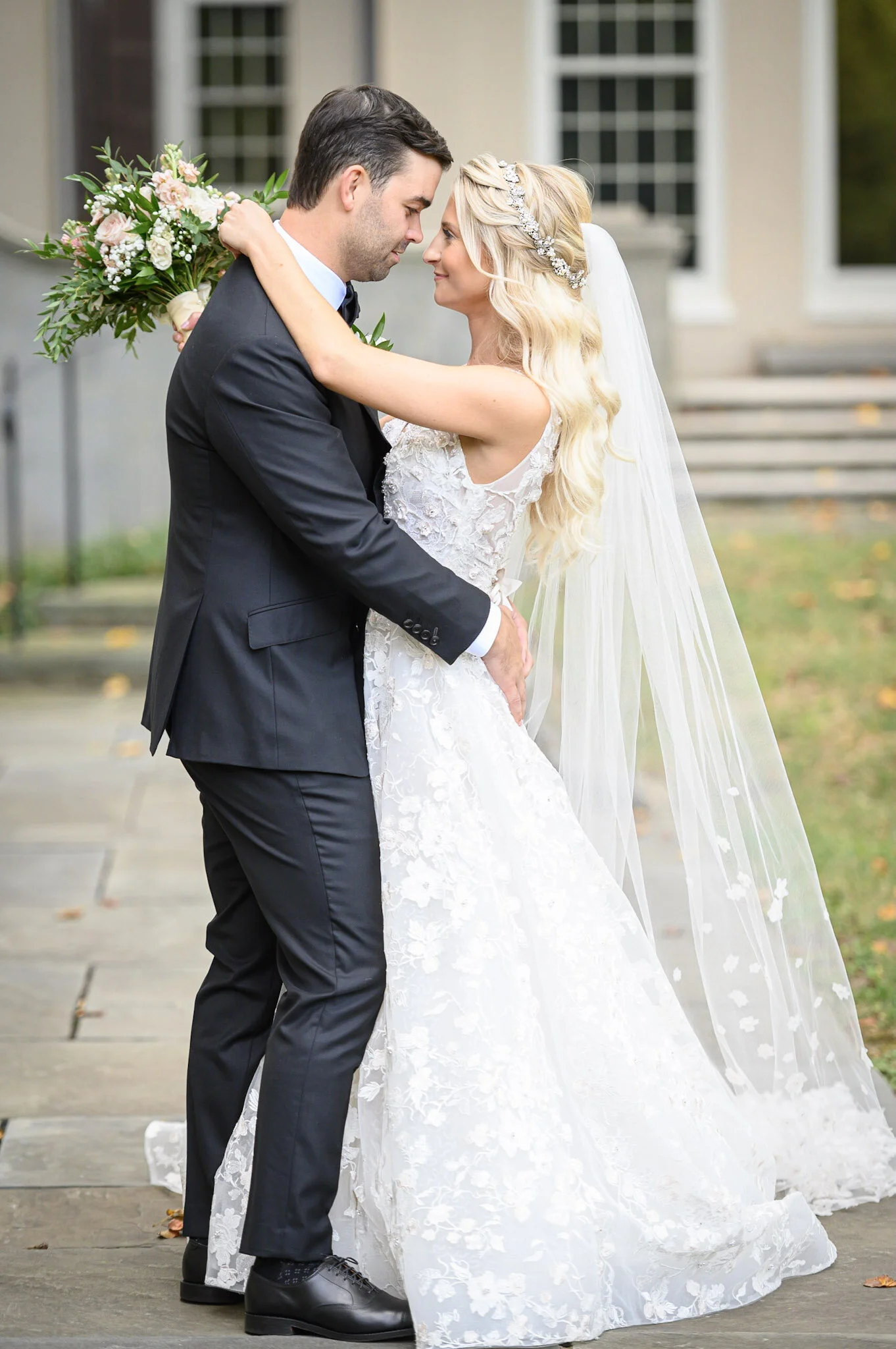 A bride and groom are standing close together outdoors, gazing into each other's eyes with their foreheads touching. The bride is wearing a white lace wedding gown with a long veil and a decorative headband, while the groom is dressed in a black tuxe