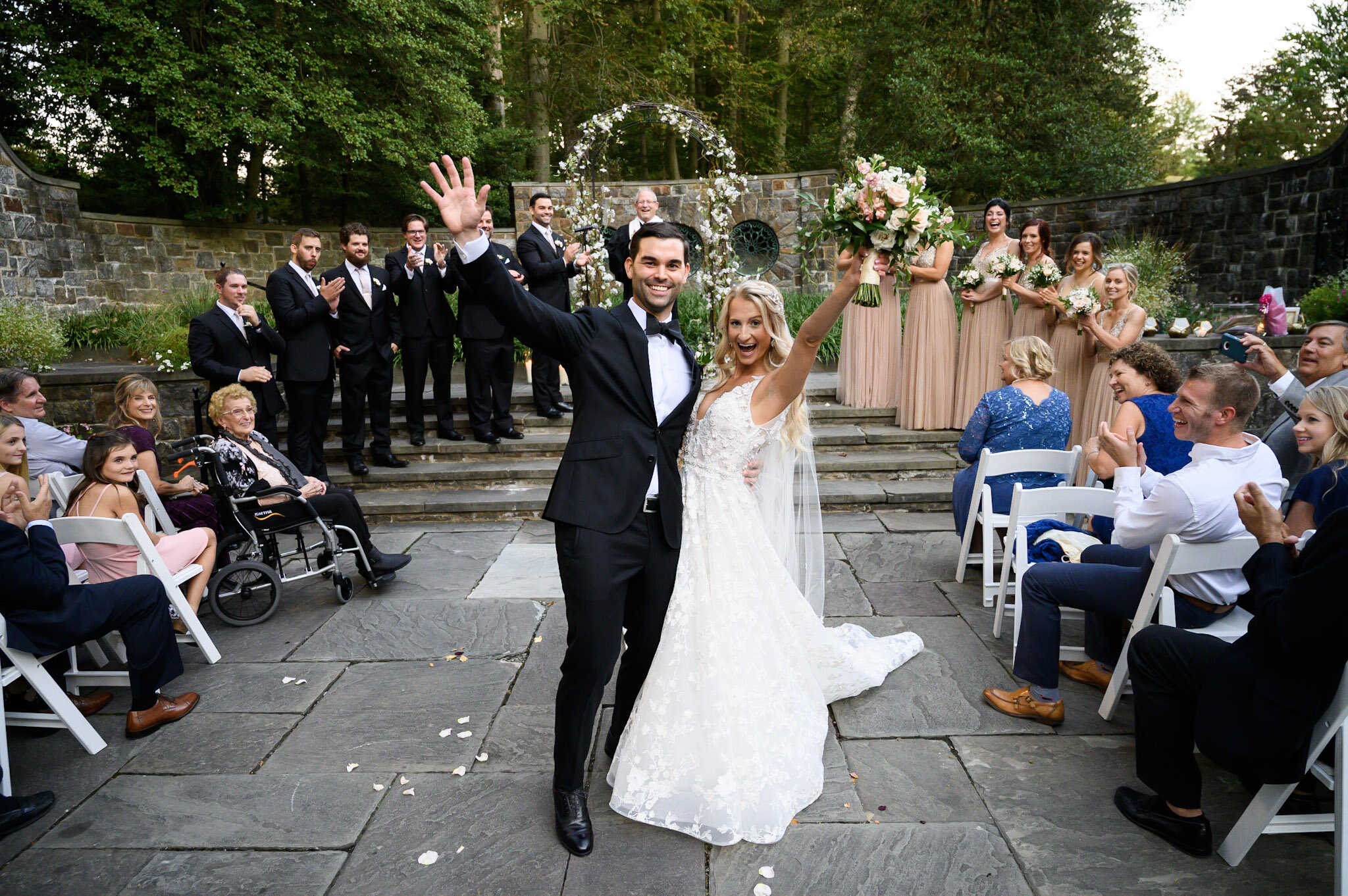 A bride and groom celebrating at their wedding ceremony outdoors, with guests seated around them, some clapping and smiling, behind a stone wall with greenery and trees.