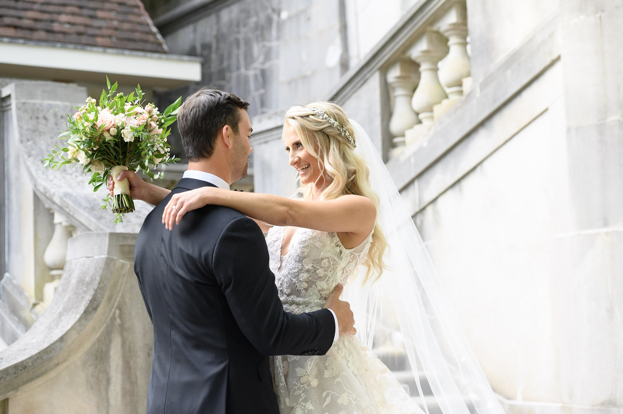 A bride and groom embrace on a staircase outside during their wedding, with the bride holding a bouquet of flowers.