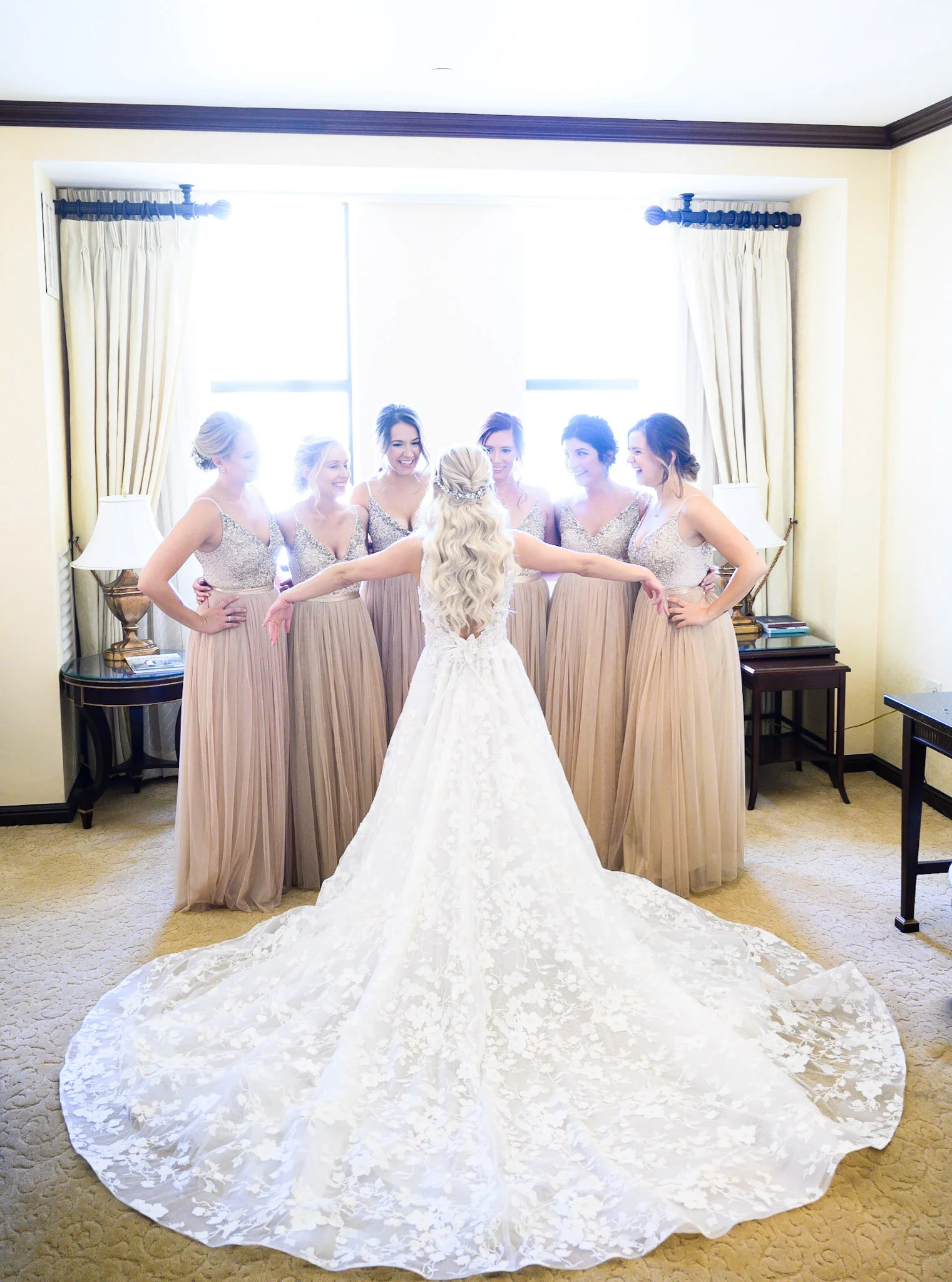 A wedding dress with a long train on a beige carpet at a wedding preparation room. Six bridesmaids in matching beige dresses are standing behind the bride, smiling and talking to each other.