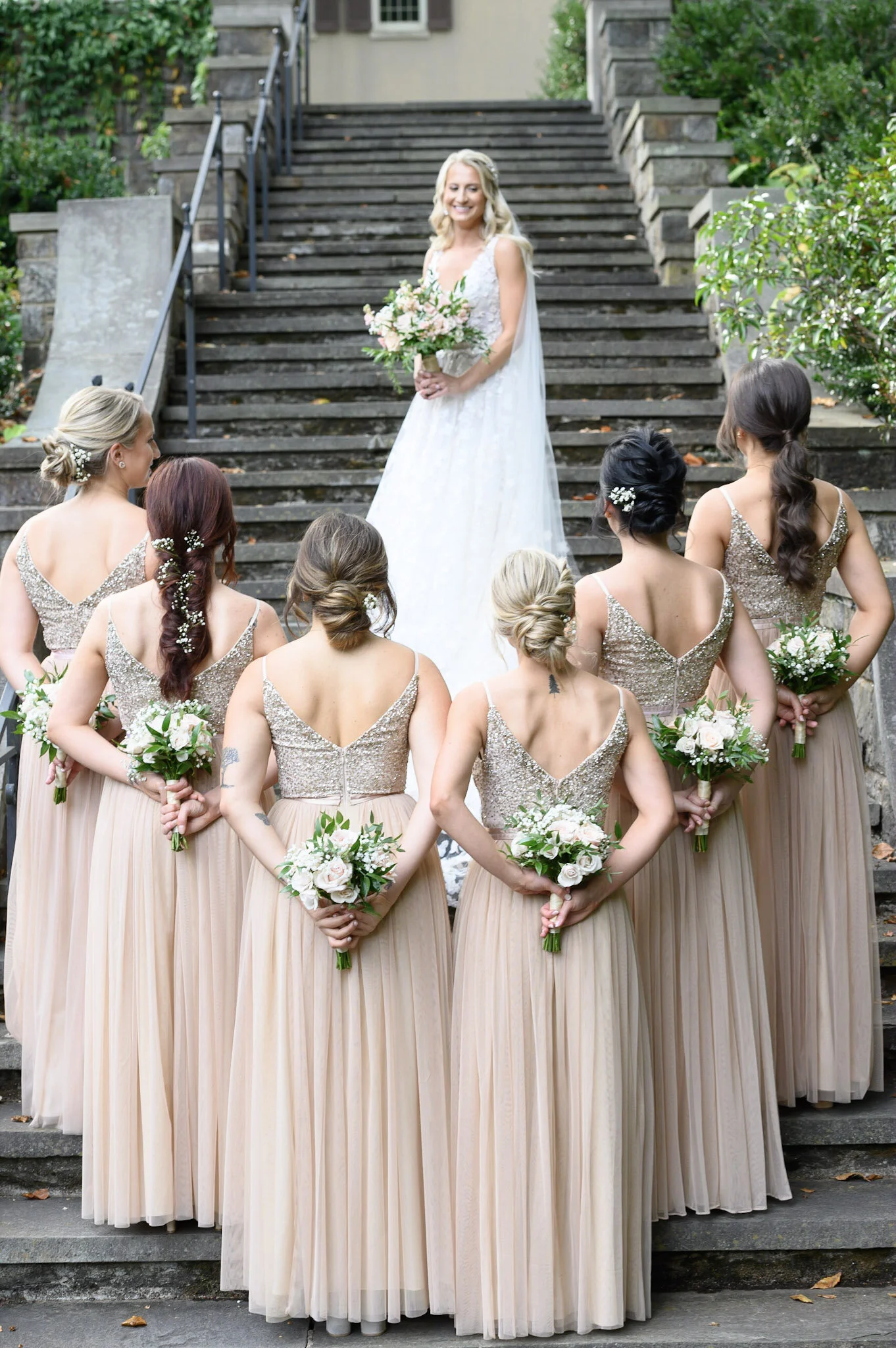 A bride holding a bouquet standing on stairs in front of bridesmaids who are dressed in long, beige and gold dresses, outdoors surrounded by green foliage.