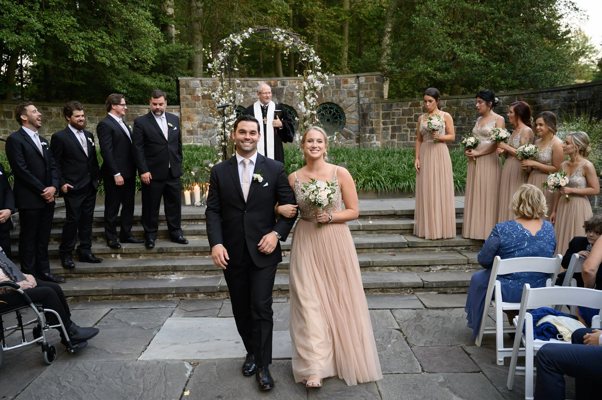 A wedding ceremony outdoors with a bride and groom walking down the steps, surrounded by bridesmaids and groomsmen, with a priest officiating, under a floral arch.
