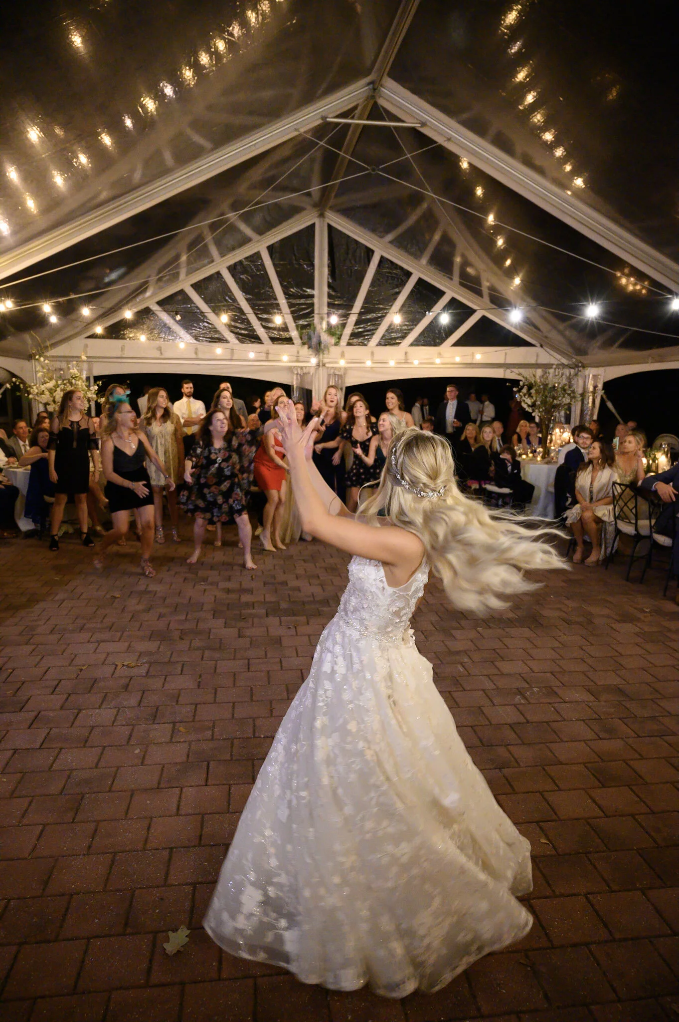 A bride with long blonde hair dancing under a clear tent decorated with string lights at night. Guests watch in the background, some standing and some seated at tables decorated with floral arrangements.