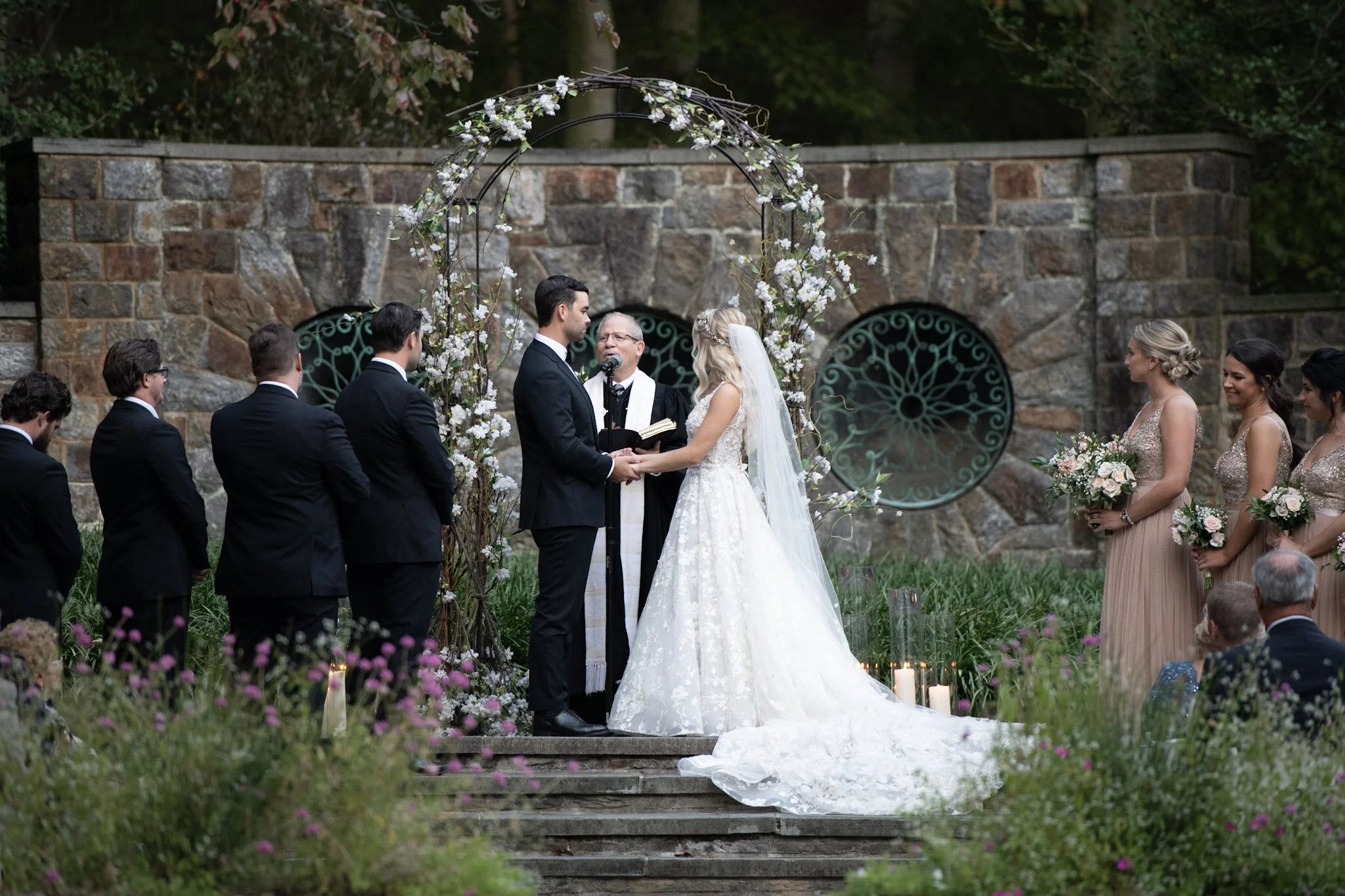 A wedding ceremony taking place outdoors with the bride and groom exchanging vows under a floral arch, surrounded by bridesmaids and groomsmen.