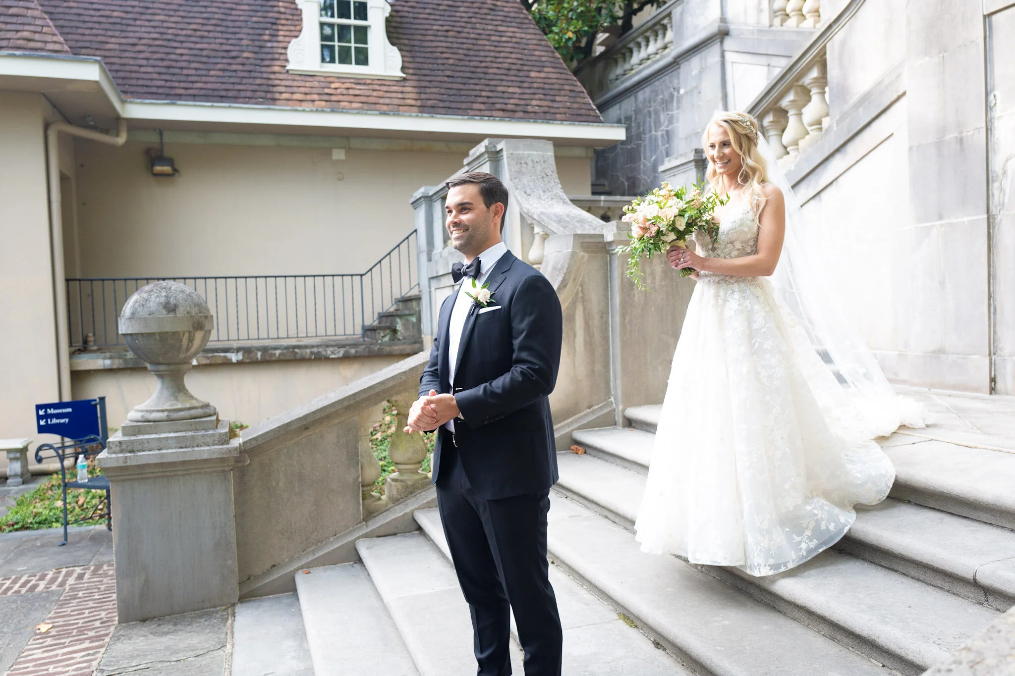 A groom and bride on stairs outside a building during their wedding, with the groom in a black tuxedo and the bride holding a bouquet of flowers.