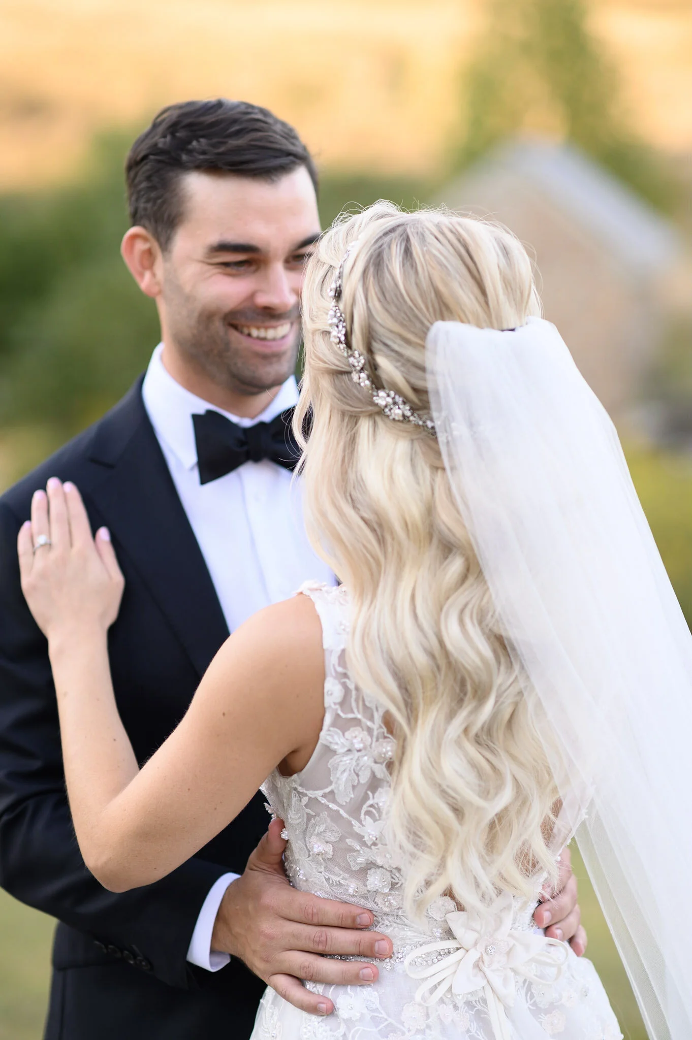 A groom in a tuxedo holding his bride in a lace wedding dress in an outdoor setting.