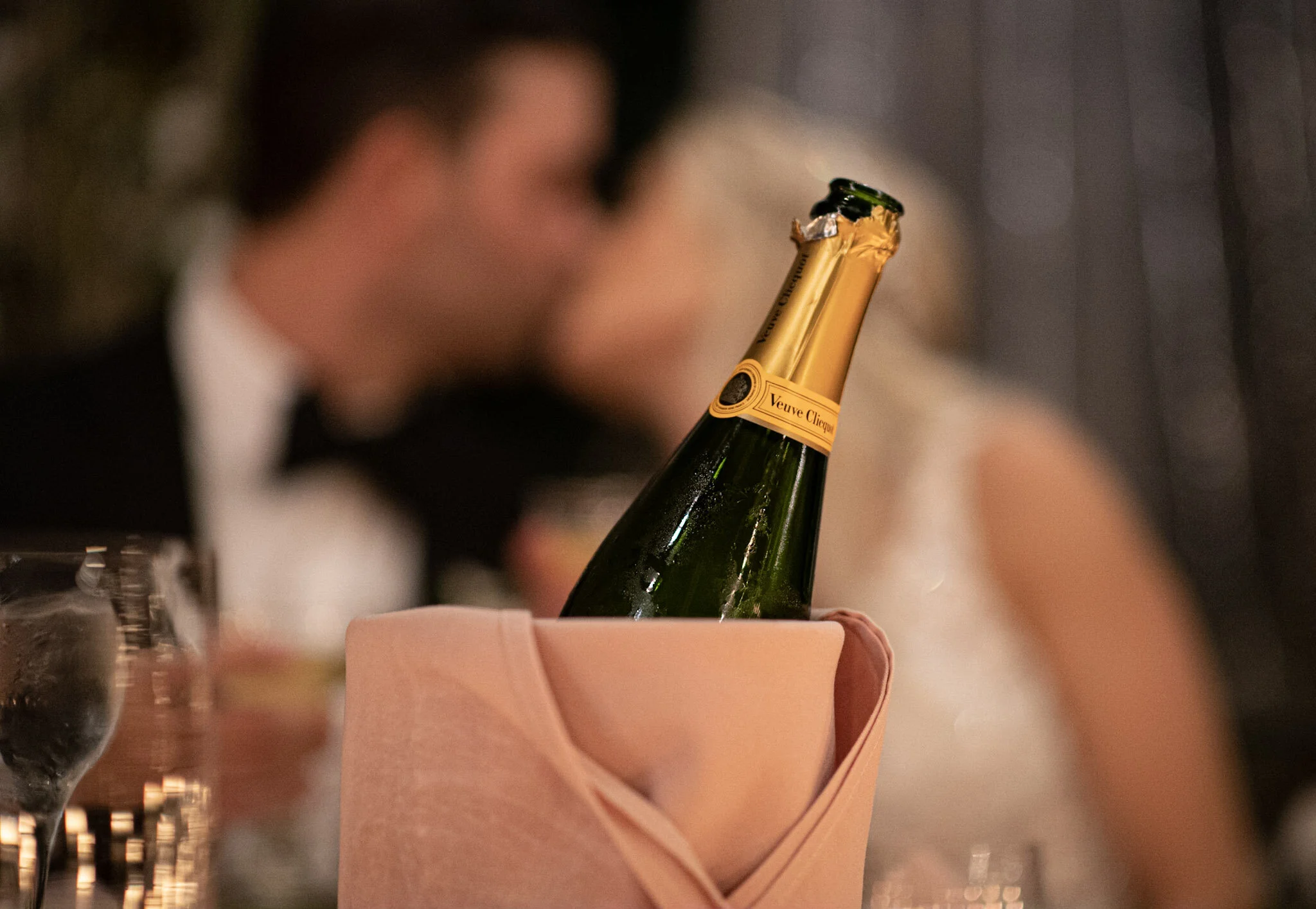 Close-up of a person holding a champagne bottle with a gold foil top, against a blurred background of a wine glass and a person's arm in a setting that appears to be a celebration or dinner.