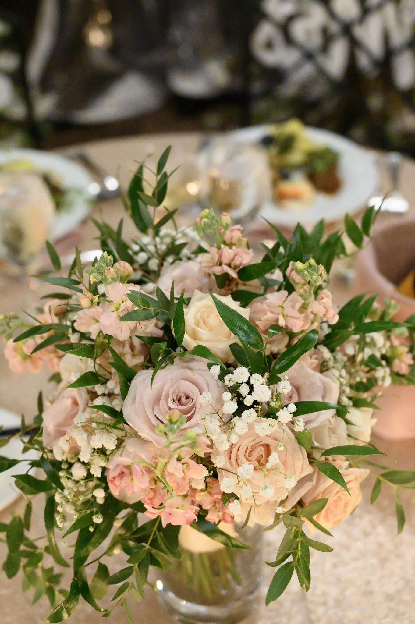 A bouquet of pink and white flowers, including roses and small white blossoms, with green leaves, in a glass vase on a table. Blurred dinner setting with plates and glasses in the background.