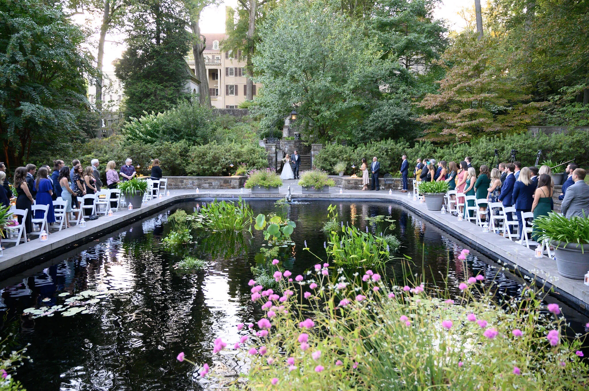A wedding ceremony taking place outdoors beside a pond, with rows of white chairs set up and guests standing on either side, surrounded by lush green trees and a historic multi-story building in the background.