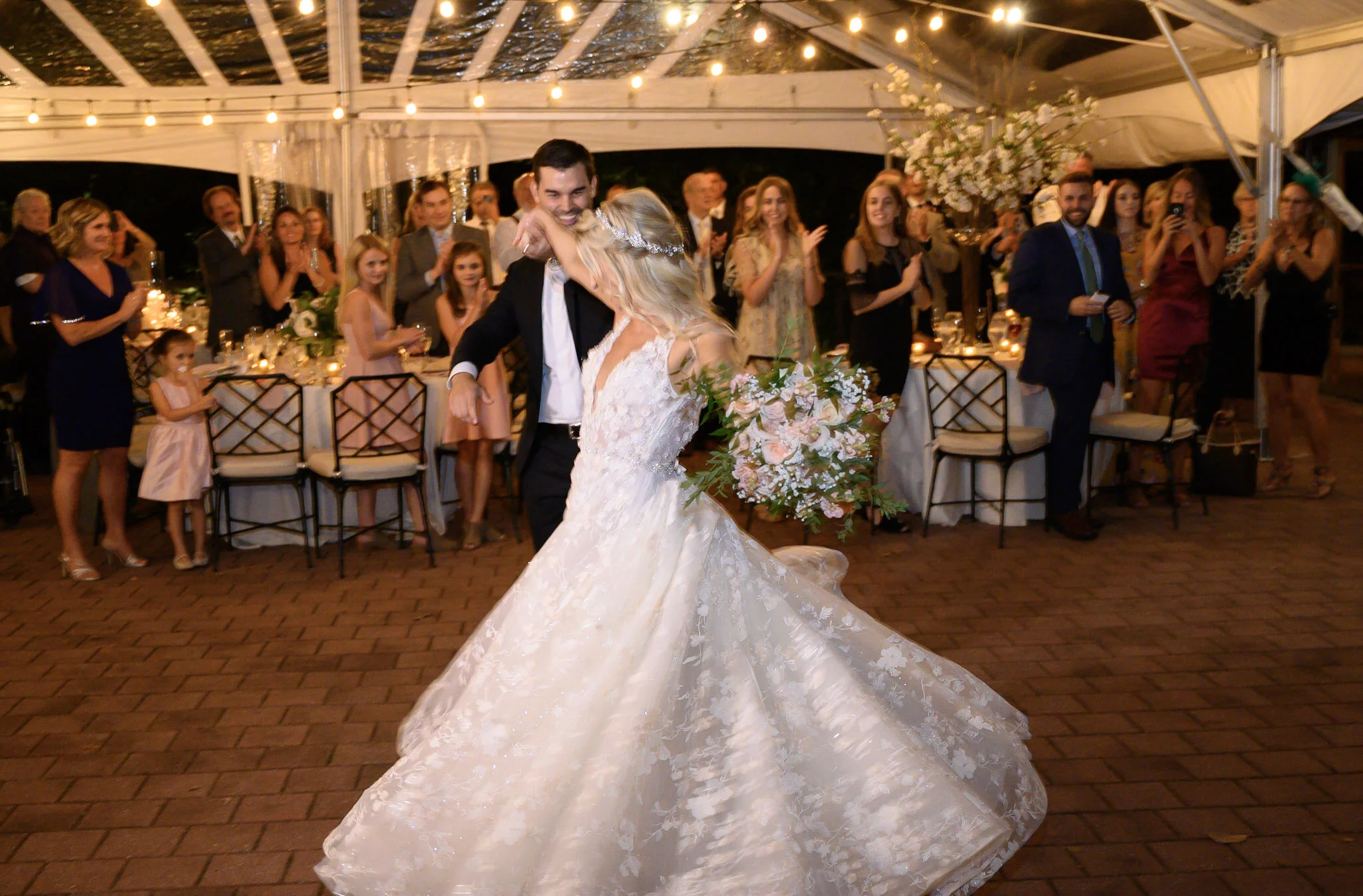 A newlywed couple dancing at their wedding reception under string lights, with guests clapping and smiling around them.
