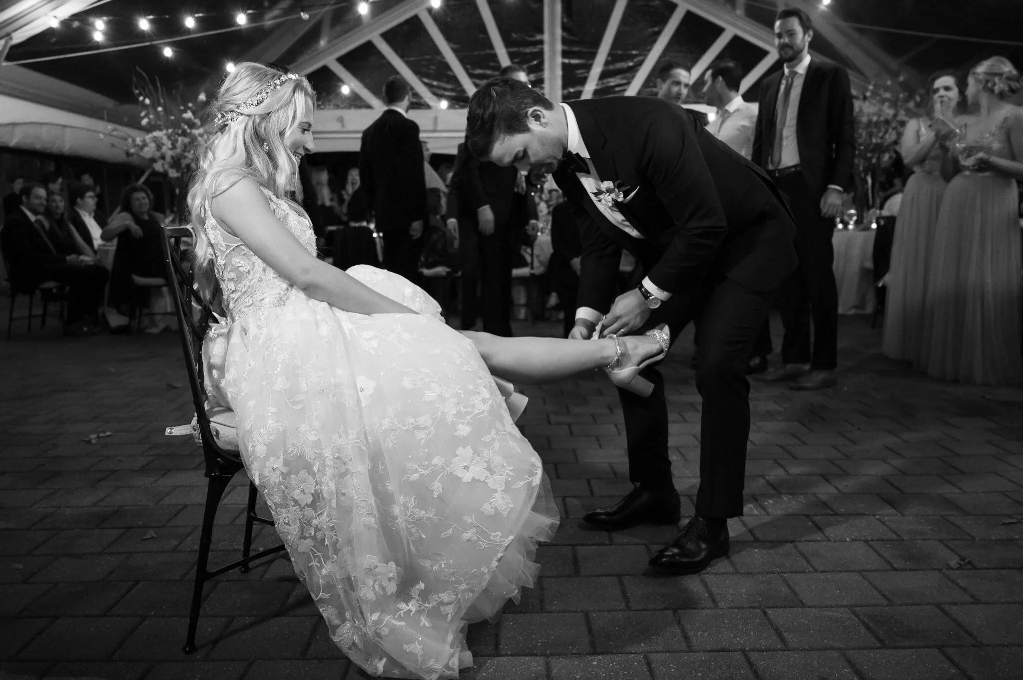 A black-and-white photo of a wedding celebration. The bride is seated on a chair, smiling at the groom, who is bending down tying her shoe. The bride is wearing a detailed lace wedding dress and a floral headband, while the groom is in a tuxedo with 