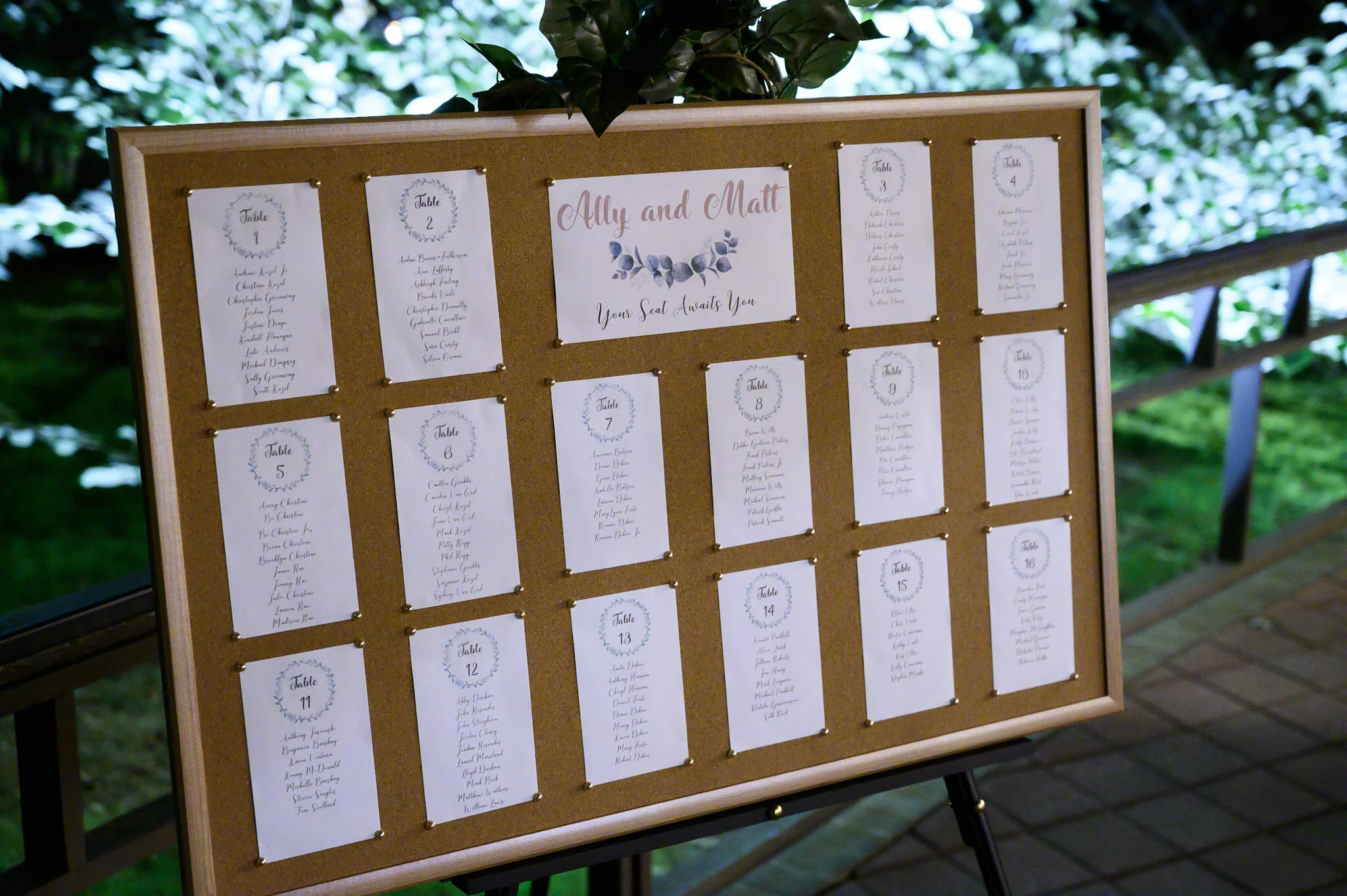 A corkboard with seating arrangements for an event, featuring white sheets with handwritten names and table numbers, including a central sign with "Ally and Matt" and "Your Seat Awaits You".