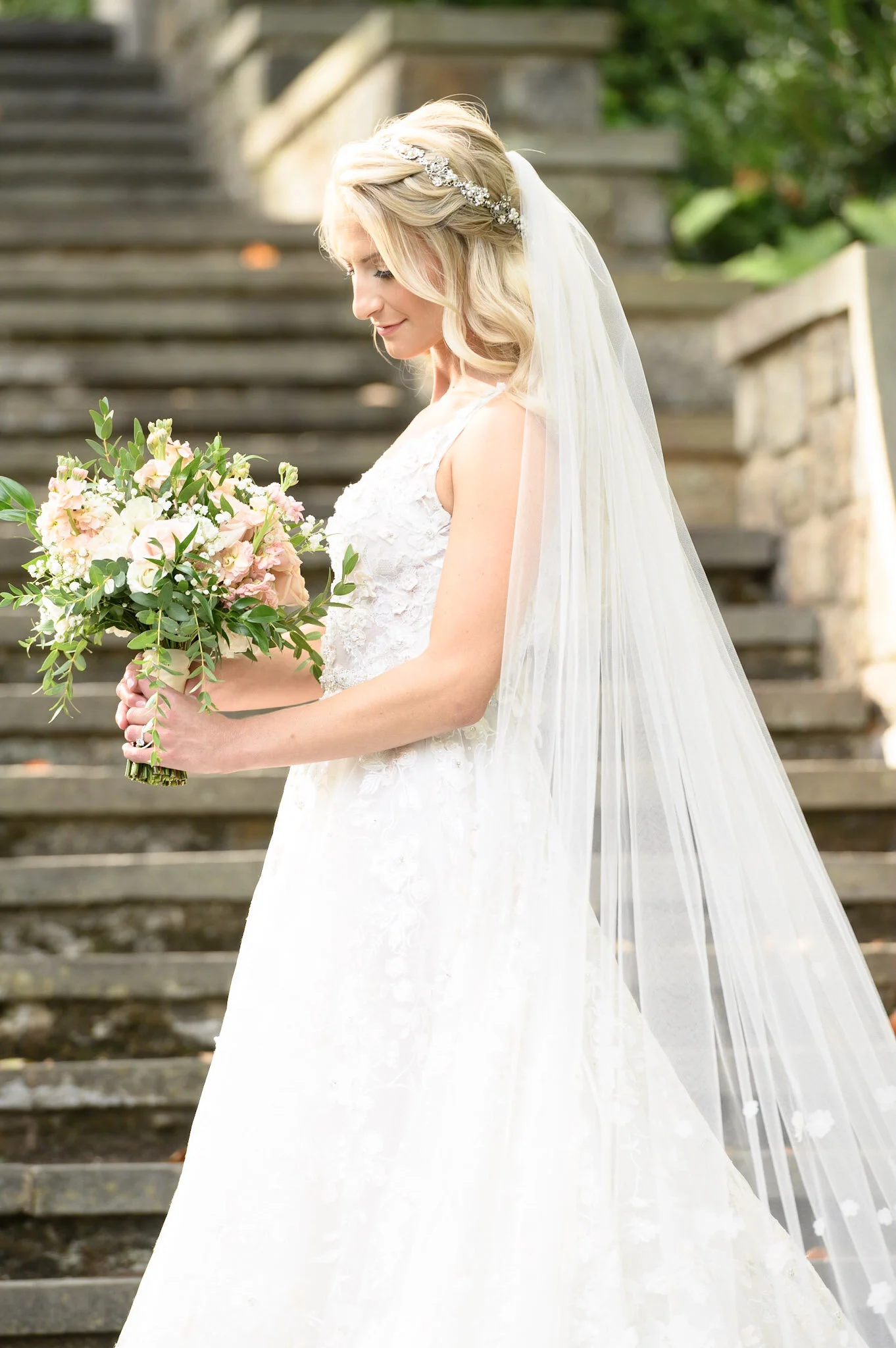A bride in a white wedding gown holding a bouquet of pink and white flowers, standing outdoors on stone steps with greenery in the background, wearing a veil and a decorative headband.
