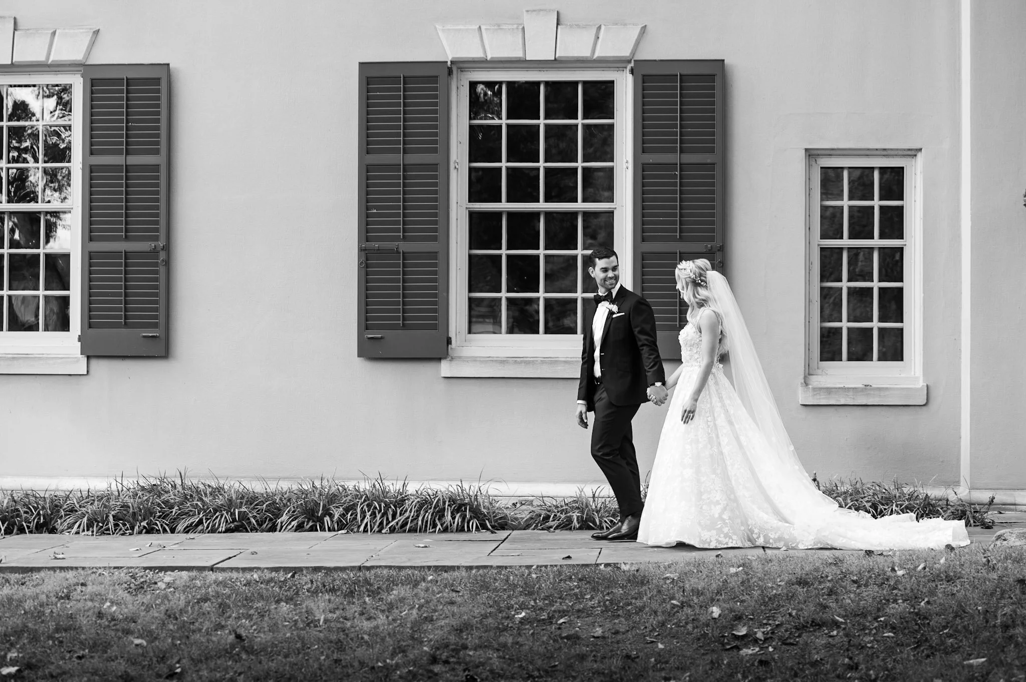 Black and white photo of a bride and groom holding hands, walking outside in front of a house with three windows and shutters.