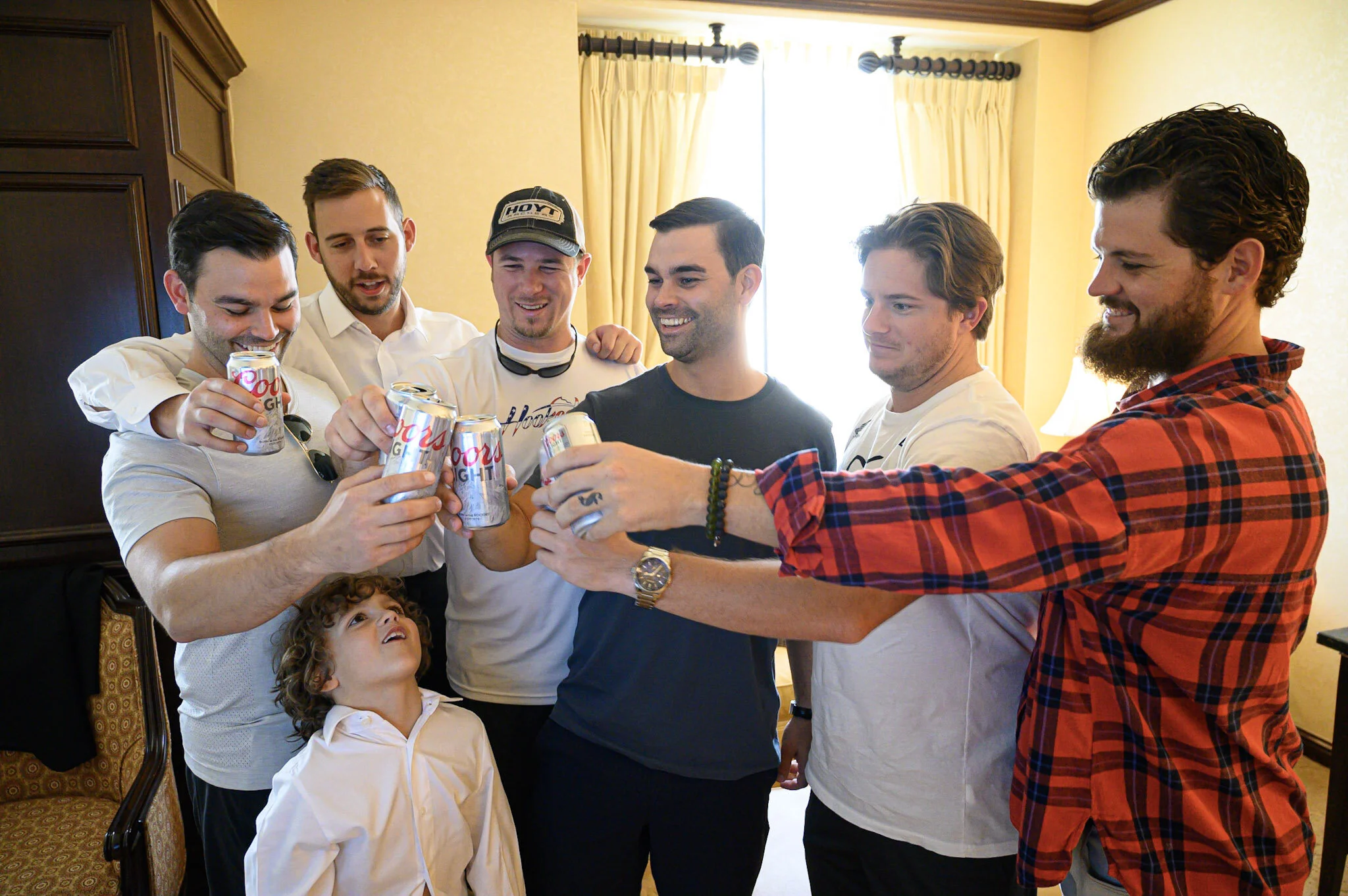 A group of six men and a young boy are gathered together, holding up cans of Coors Light beer and smiling. They are in a room with yellow walls, a window with curtains, and wooden furniture.