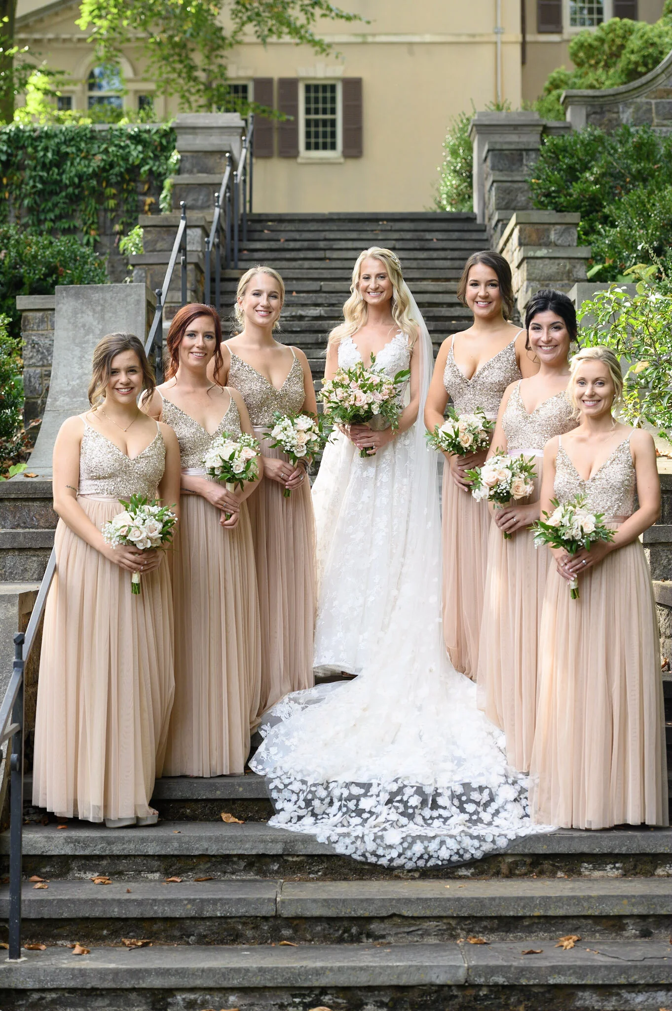 Group of women in formal dresses, including a bride in a white wedding gown, standing on steps outside a building with green foliage.