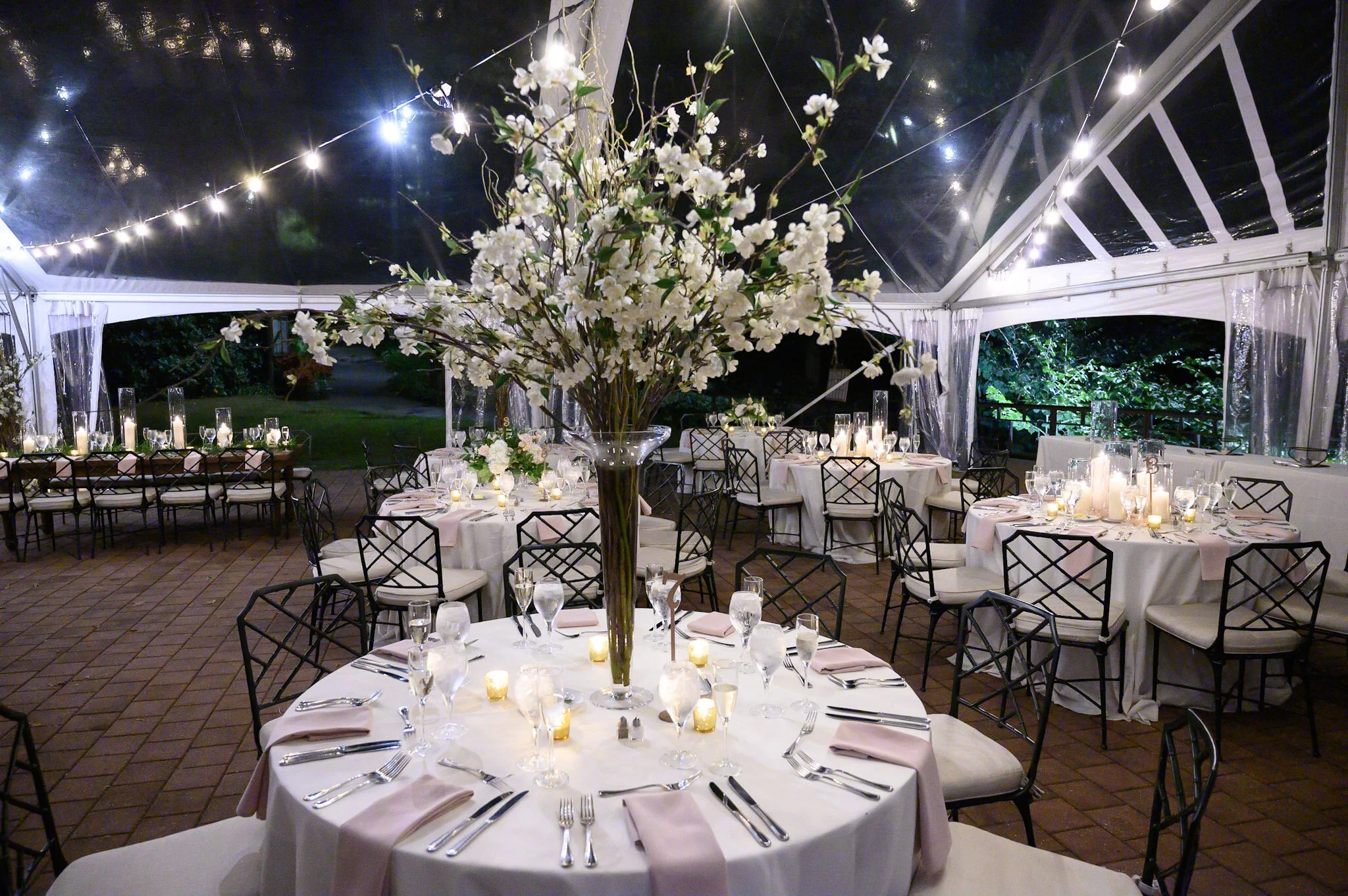 Elegant outdoor banquet setup inside a clear tent at night, featuring round tables with white tablecloths, pink cloth napkins, wine glasses, and cutlery. The centerpiece is a large floral arrangement of white flowers, and string lights hang across th