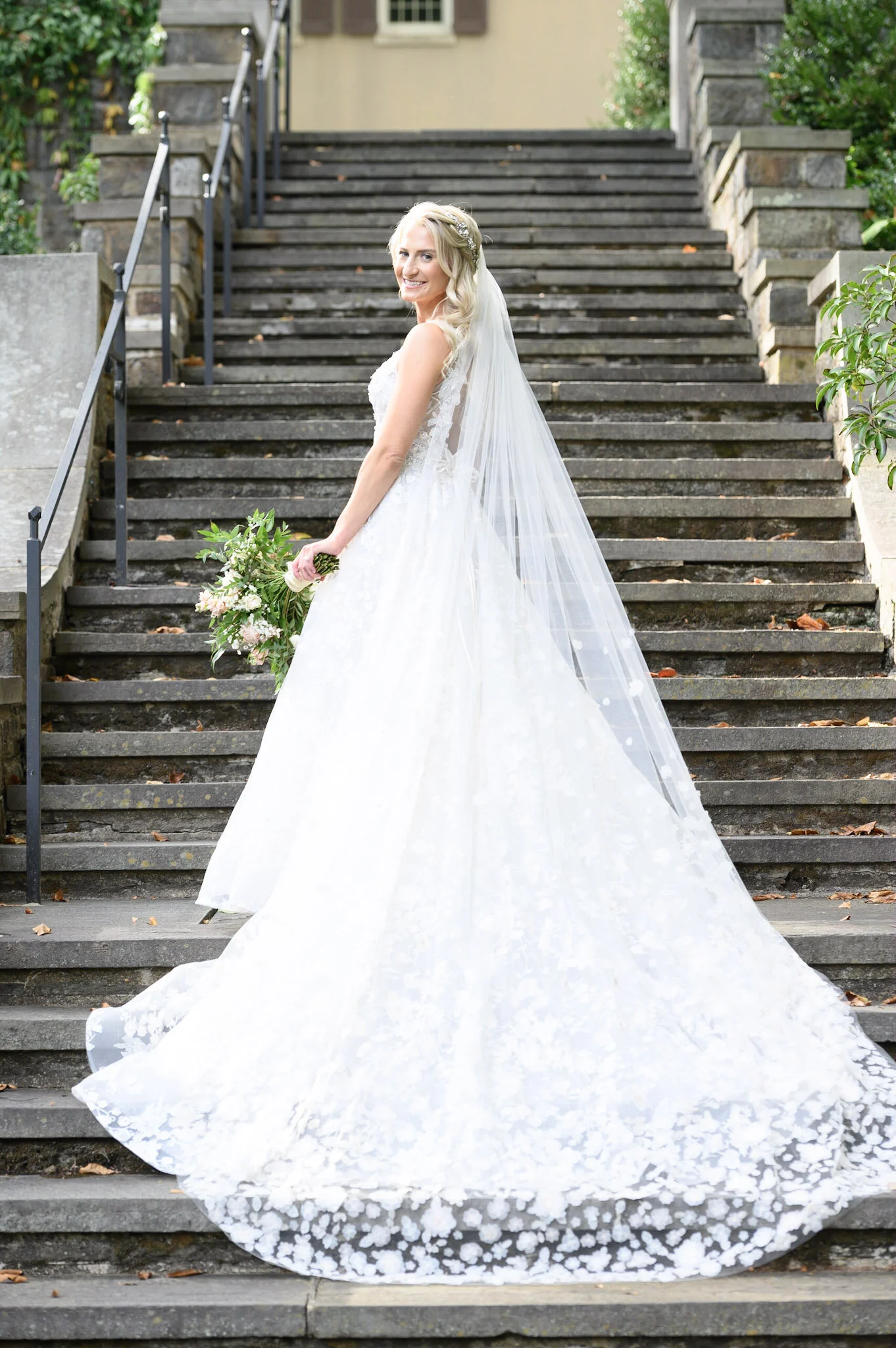 A smiling blonde bride in a white wedding gown holding a bouquet of flowers, standing on outdoor stone stairs surrounded by greenery.