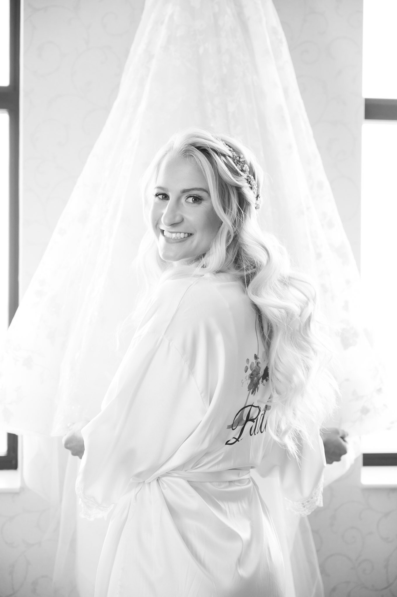 Black and white photograph of a woman with blonde hair styled in loose waves, wearing a floral headband and a satin robe with the word 'Bride' written on the back, standing in front of a window with sheer curtains.