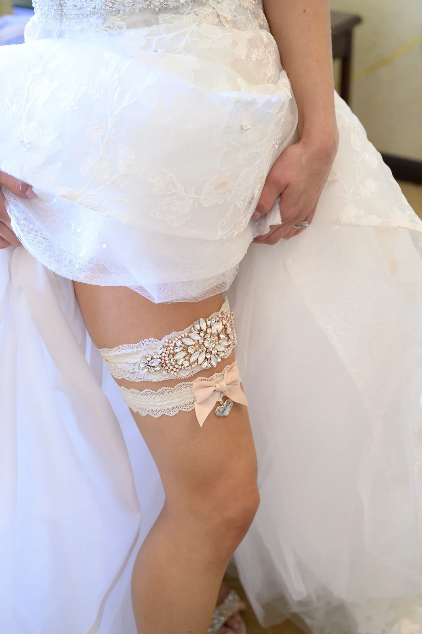 Close-up of a bride wearing a white wedding dress with lace and floral embroidery, lifting her dress to reveal lace garter with a large jewel embellishment and a small bow, while holding her dress with her other hand.