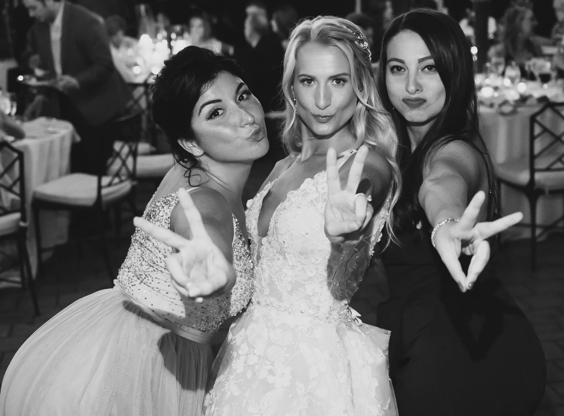 Three women taking a selfie at a wedding reception, smiling and making peace signs with their hands, with blurred tables and guests in the background.