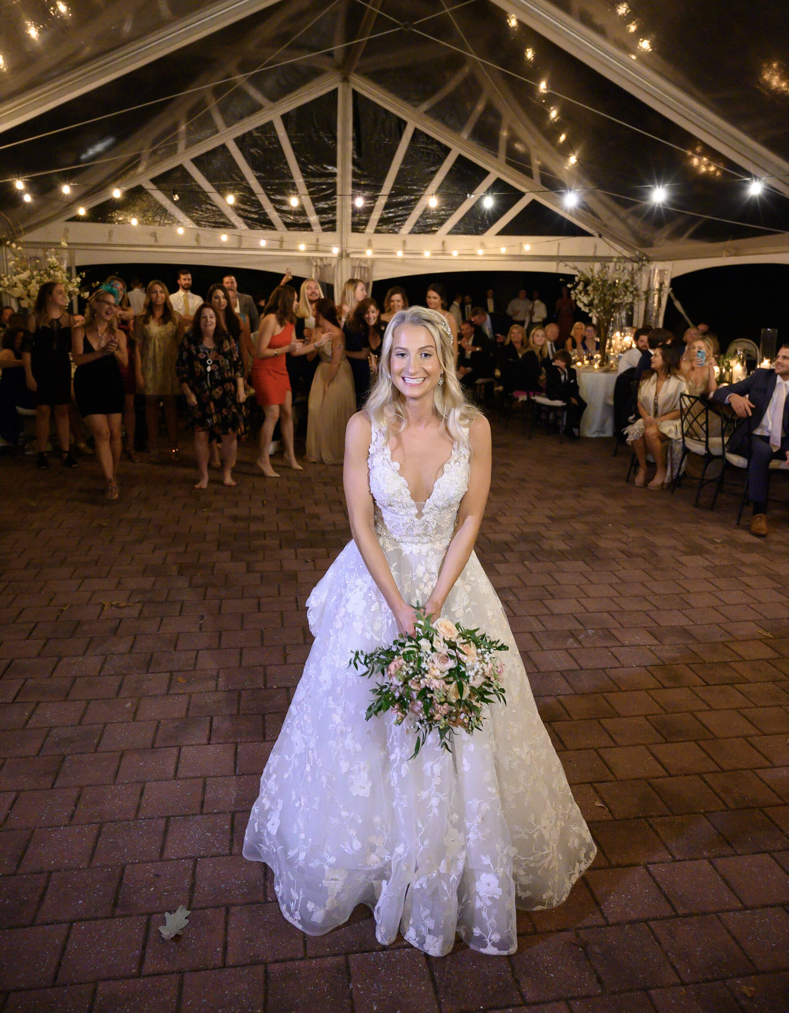 A bride in a white lace wedding dress is smiling and holding a bouquet of flowers at a wedding reception under a large illuminated tent with guests in the background.