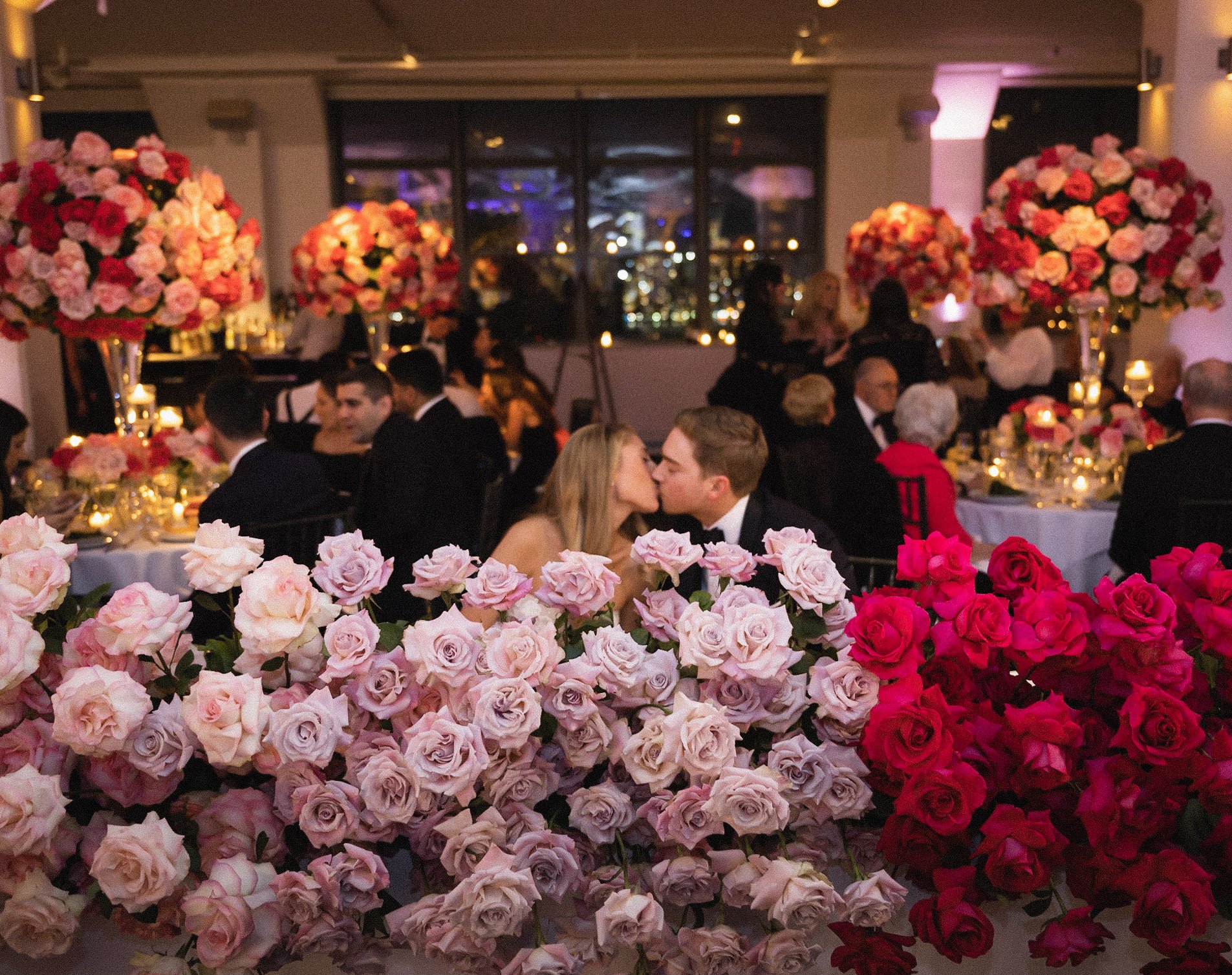 An elegant indoor wedding reception with a large floral centerpiece in shades of pink and red roses in the foreground, and guests seated at tables in formal attire. The background features a window with city lights outside.