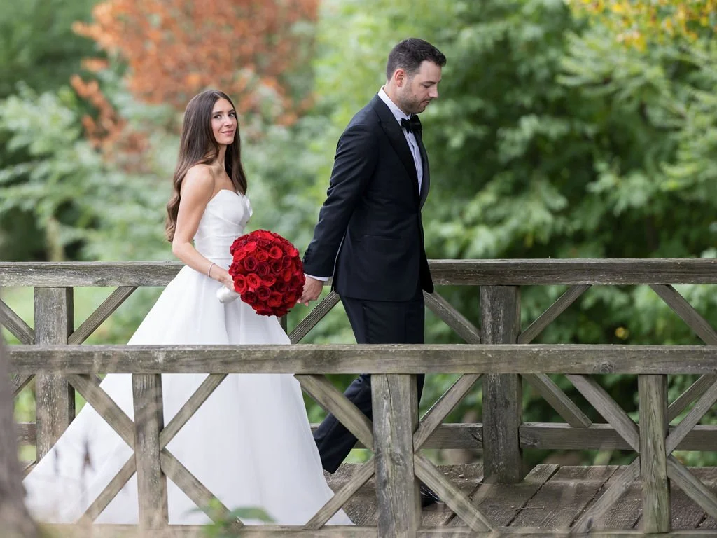 Bride in white wedding gown holding a bouquet of red roses, walking behind groom in a black tuxedo on a wooden bridge in a lush green outdoor setting.