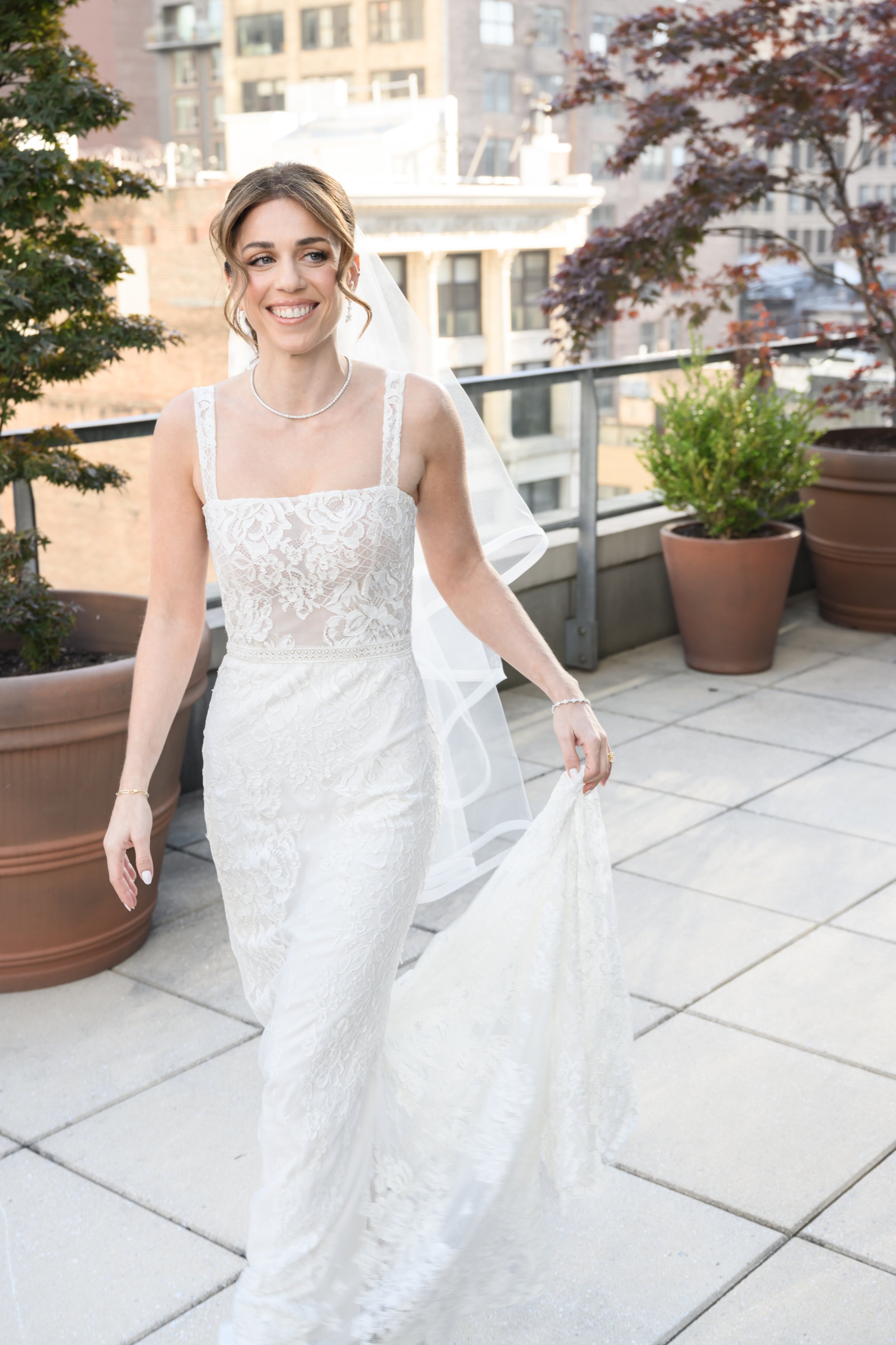 A bride in a white lace wedding dress, smiling, on a rooftop terrace with potted plants and city buildings in the background.