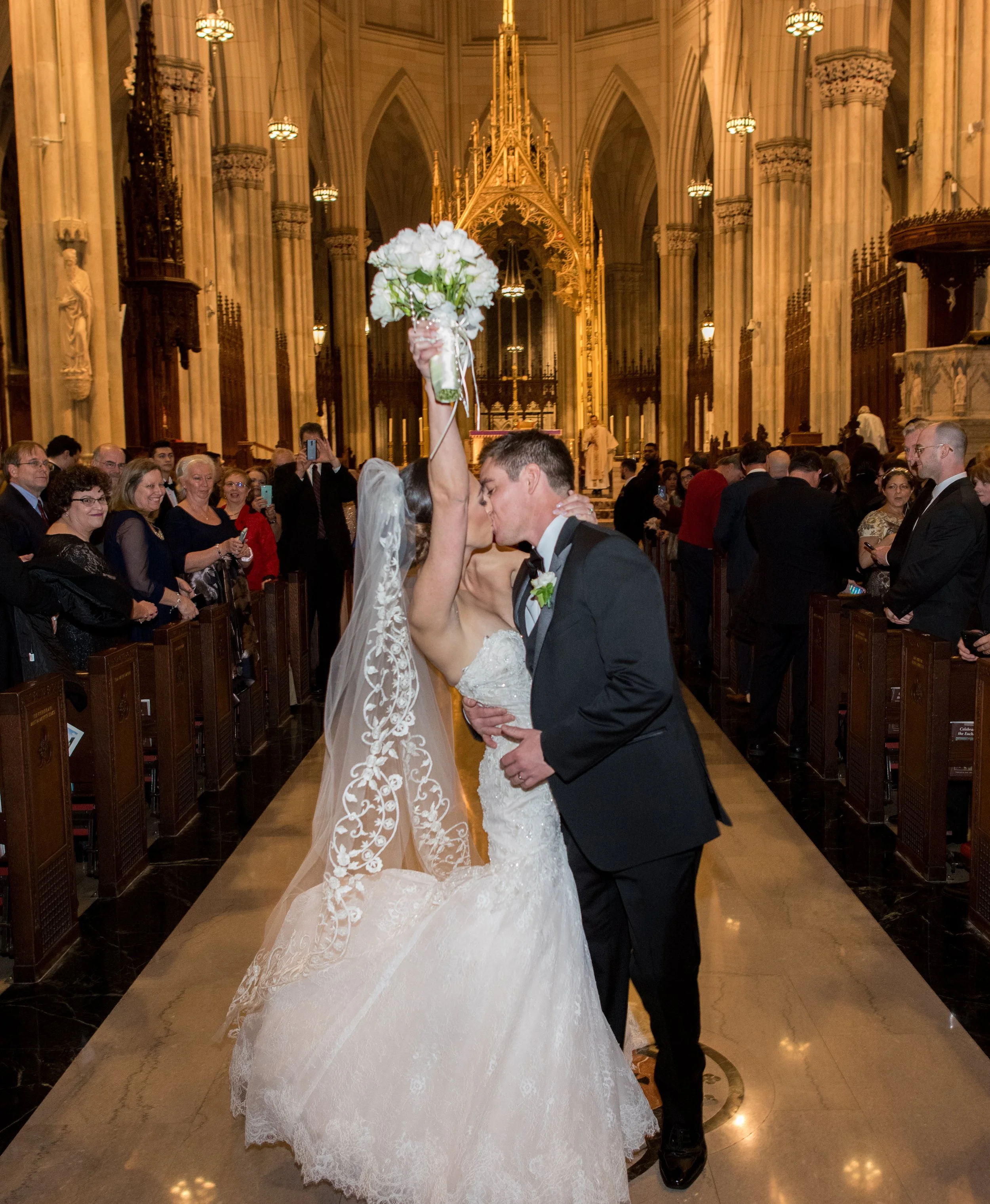 A bride and groom kissing during their wedding ceremony inside a church, with a woman holding a bouquet of white flowers above them, and guests watching and taking photos.