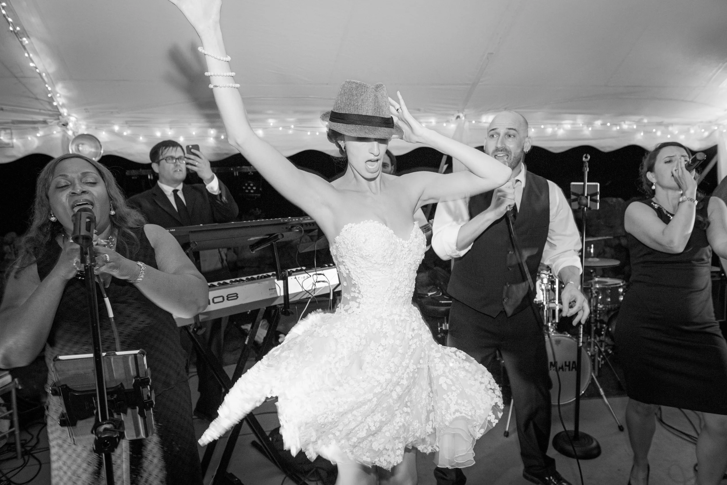 A woman in a wedding dress dancing with people playing music and singing at a celebration or wedding reception, under a tent decorated with string lights.