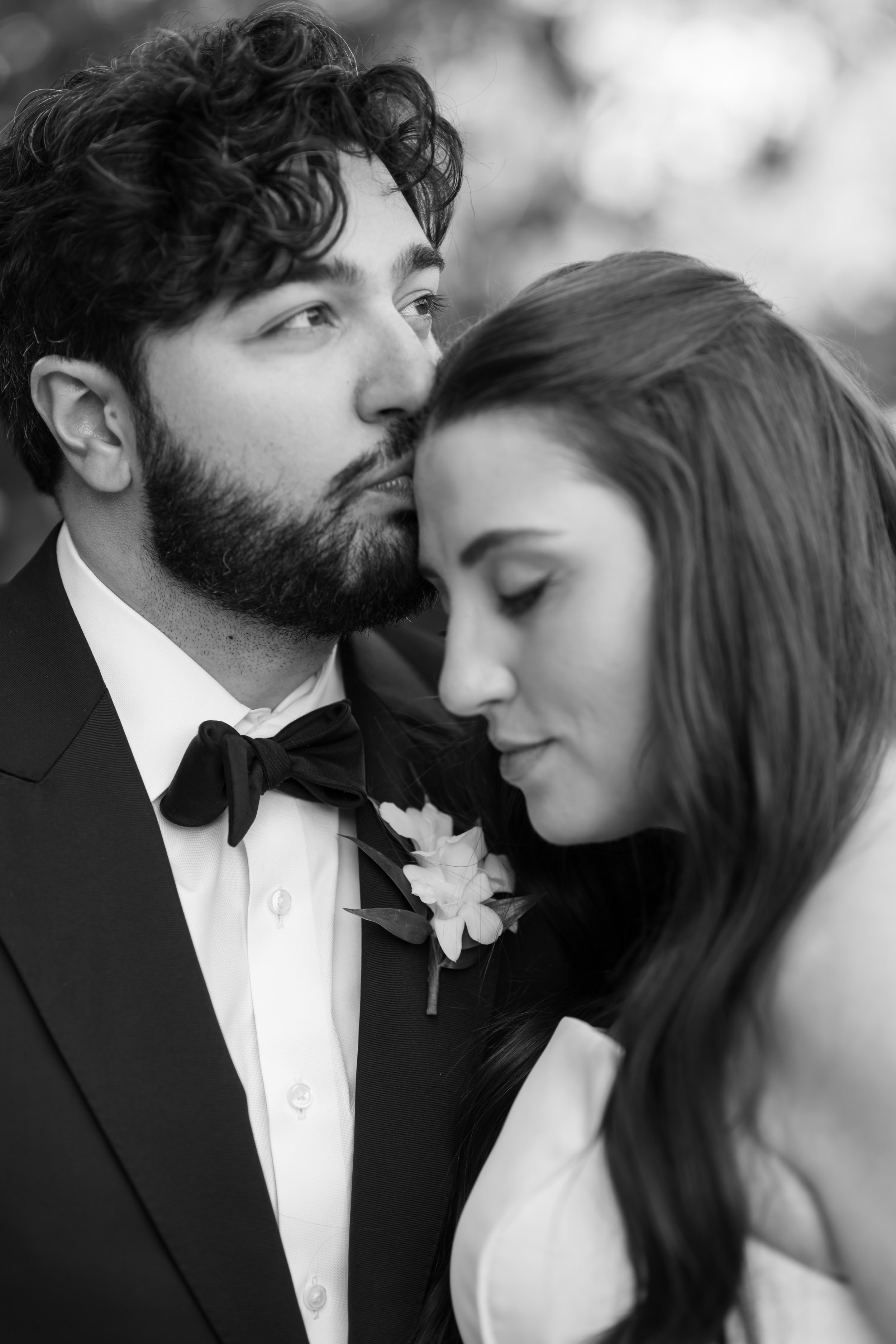 A black and white photograph of a couple at a wedding, with the man wearing a tuxedo and the woman in a white dress, their foreheads touching and eyes closed, sharing an intimate moment.