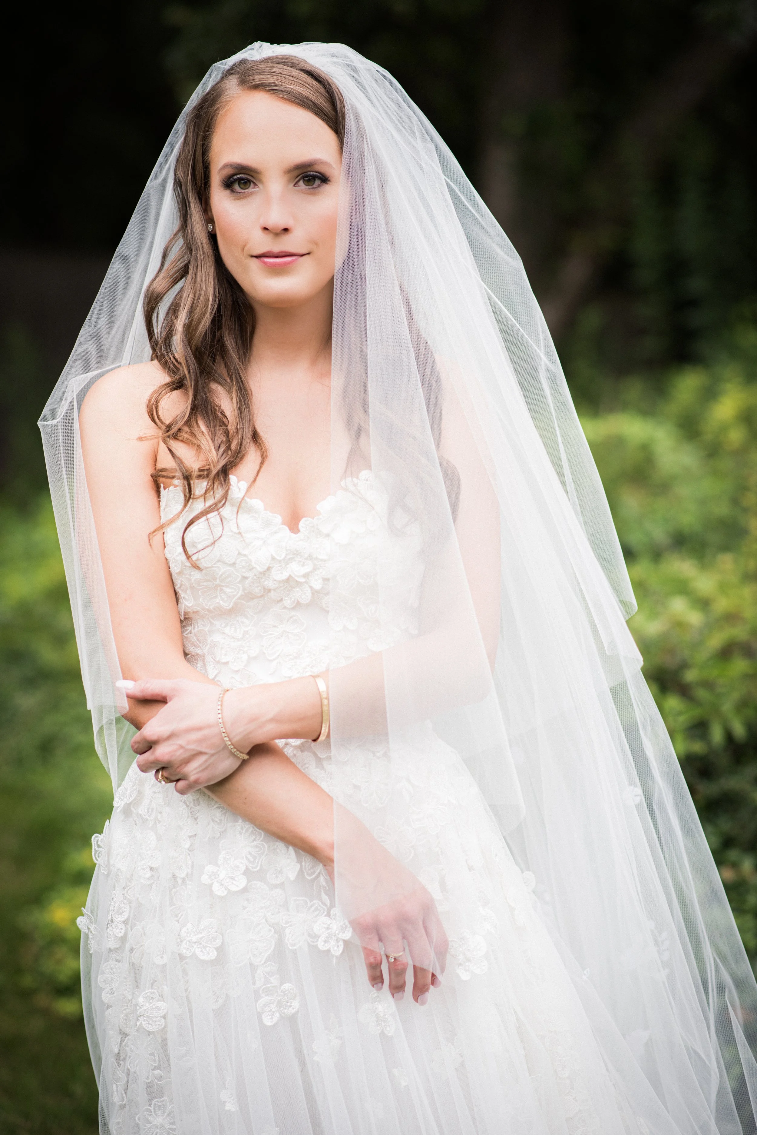A bride in a white lace wedding dress and veil standing outdoors with green foliage in the background.