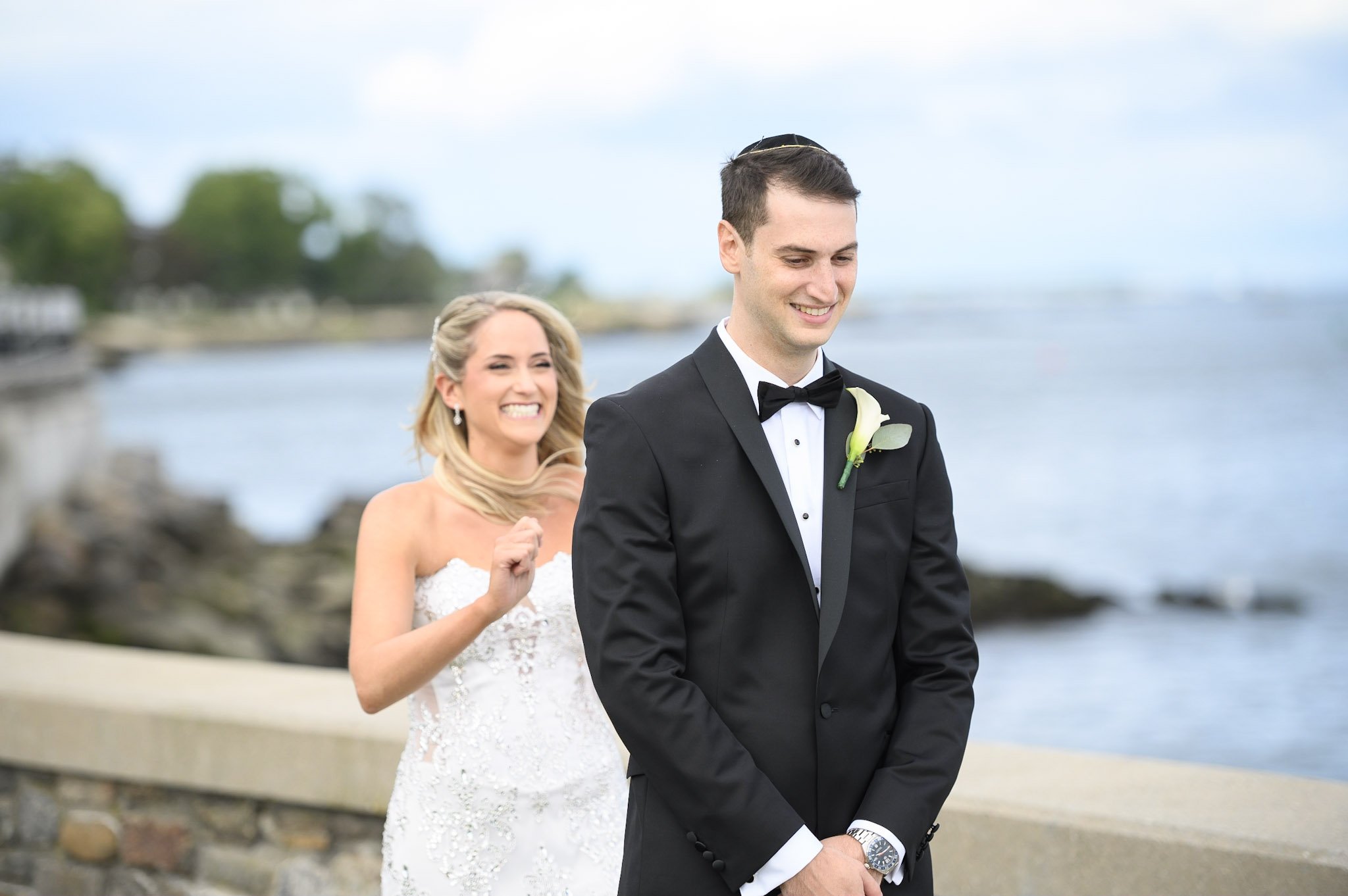 A newlywed couple standing outdoors, the bride smiling enthusiastically and the groom smiling shyly, dressed in wedding attire with a scenic ocean background.