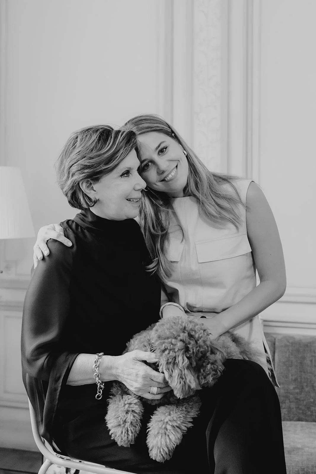 An older woman and a young woman smiling and hugging, with the older woman holding a curly-haired puppy, inside a room with decorative wall panels and a table lamp.